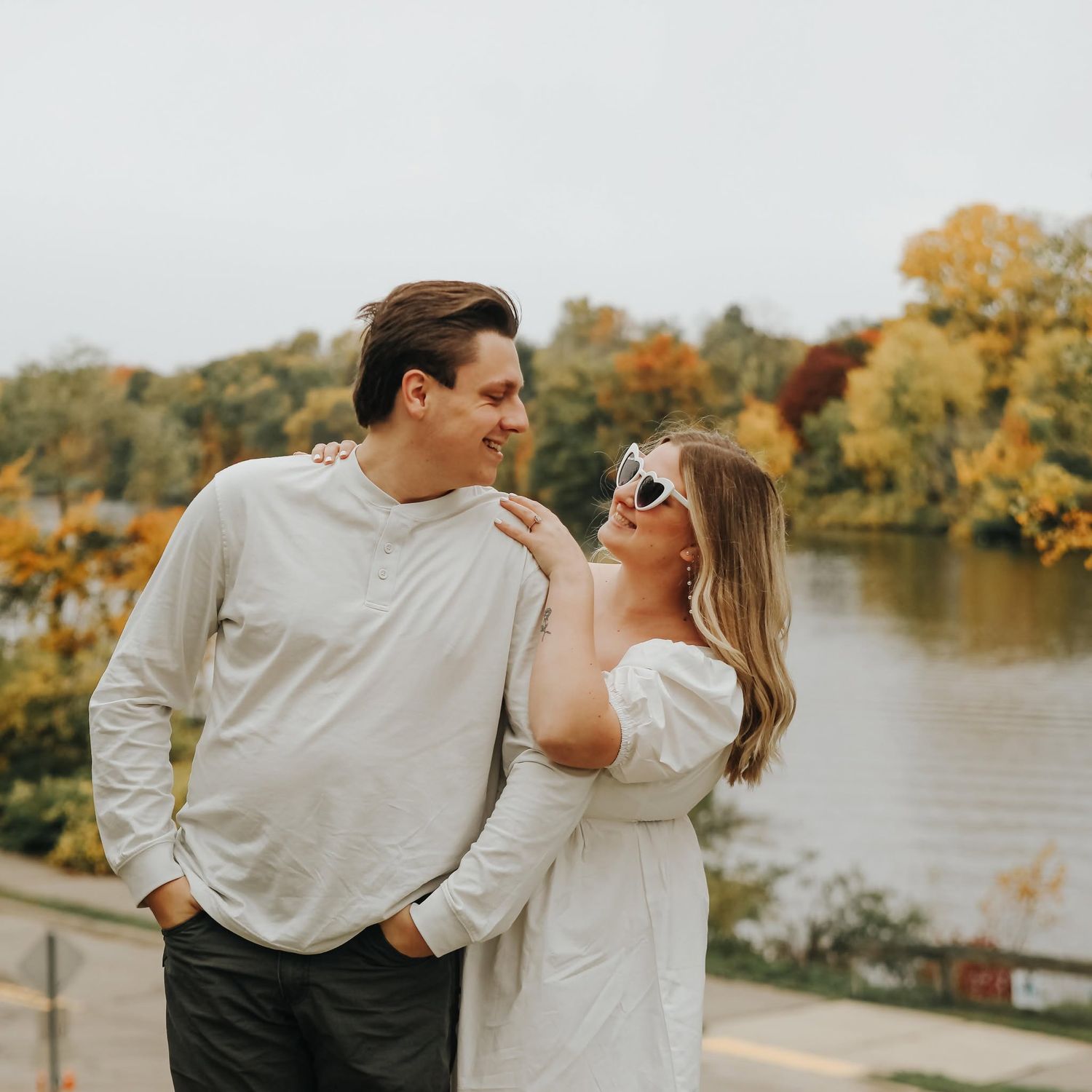 Couple posing near a lake with autumn foliage, wearing white outfits, smiling at each other outdoors.