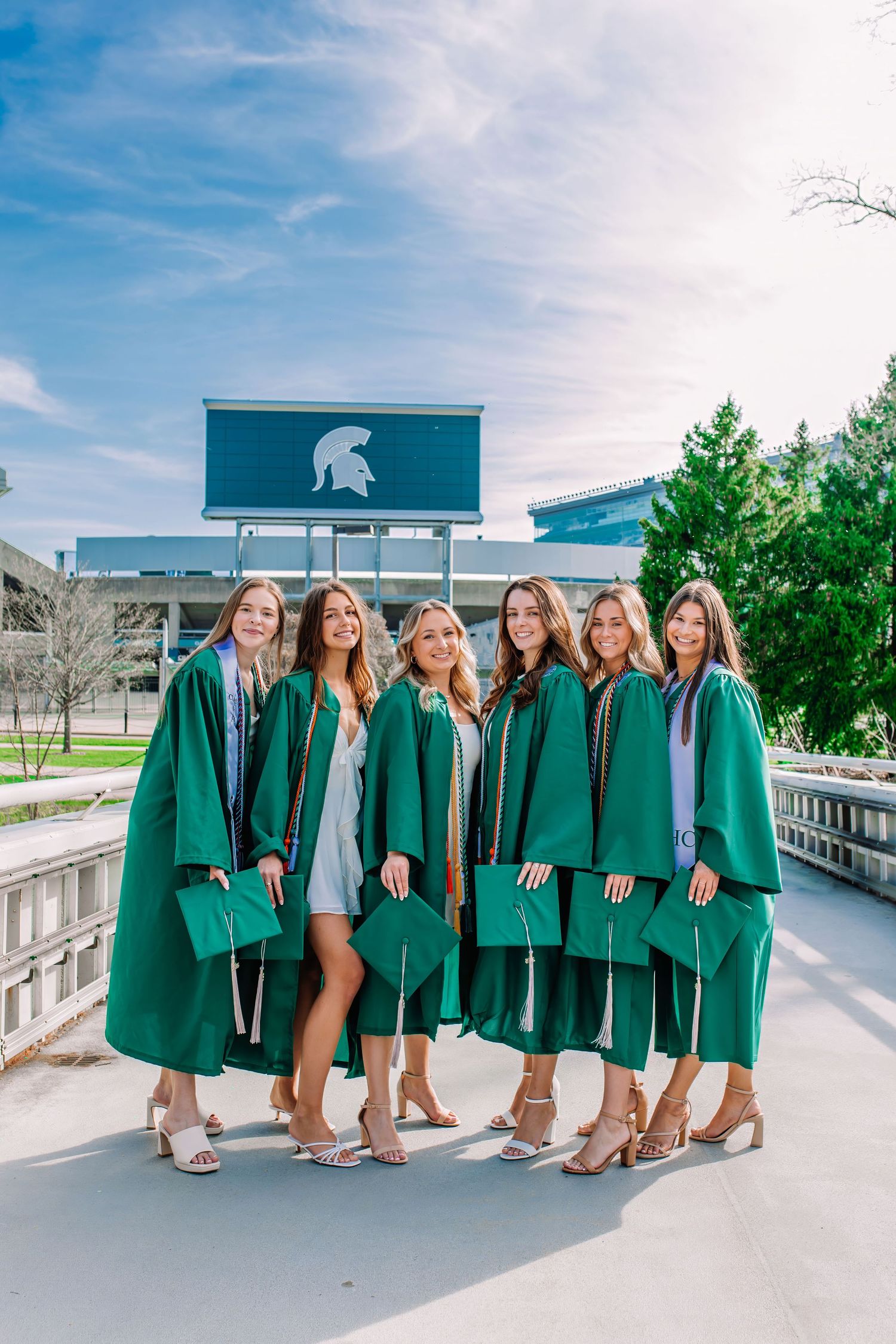 Six women in green Michigan State University graduation gowns pose together outside Spartan Stadium.