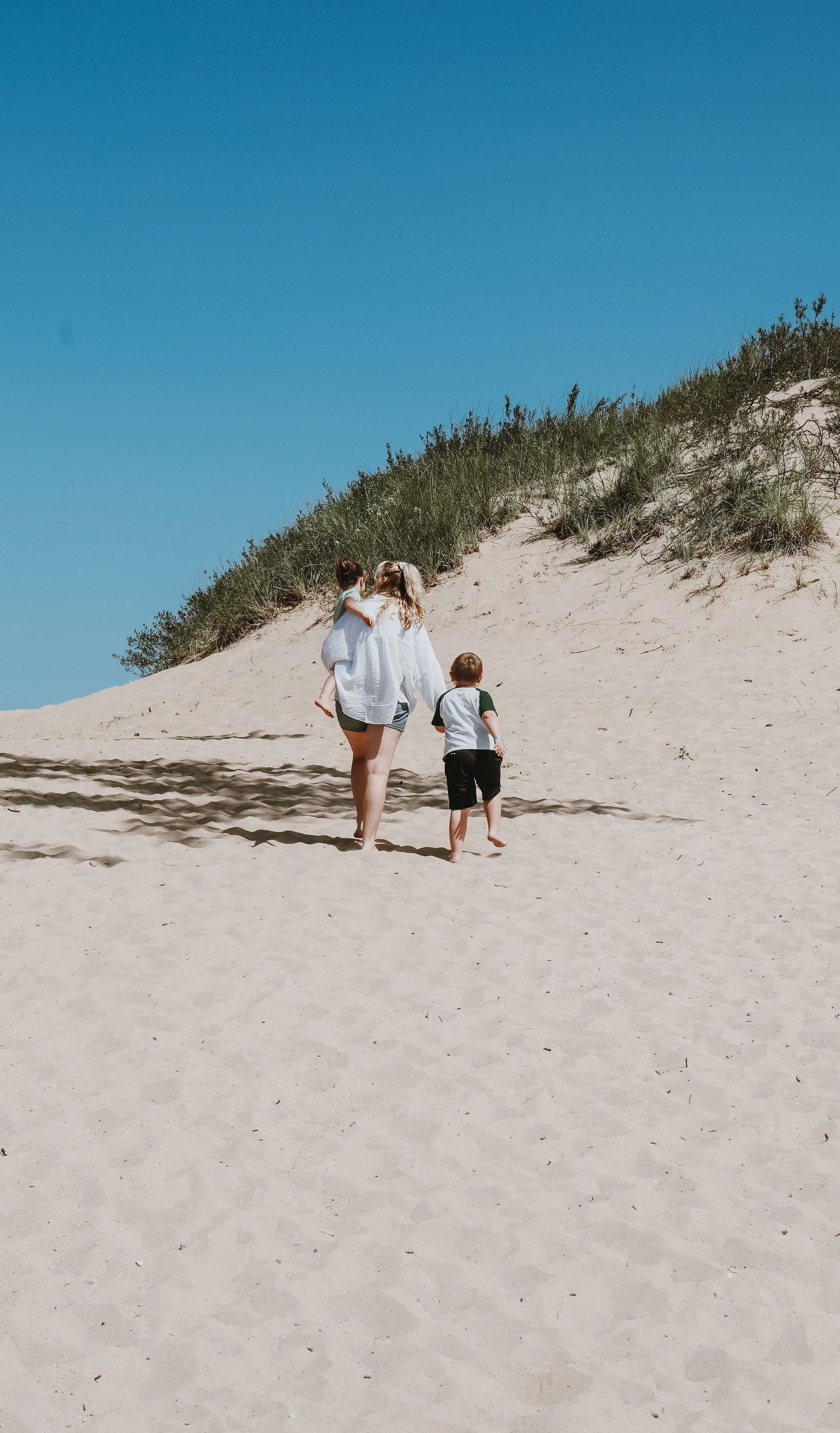 People walking up sandy dune slope against bright blue sky on sunny day.