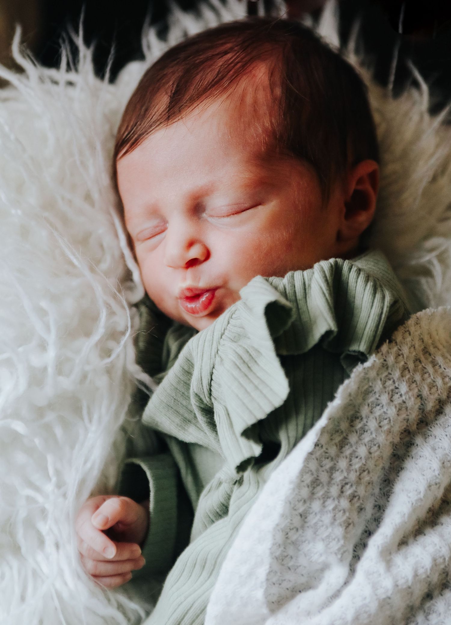 Sleeping newborn baby wearing mint green outfit on white furry blanket.