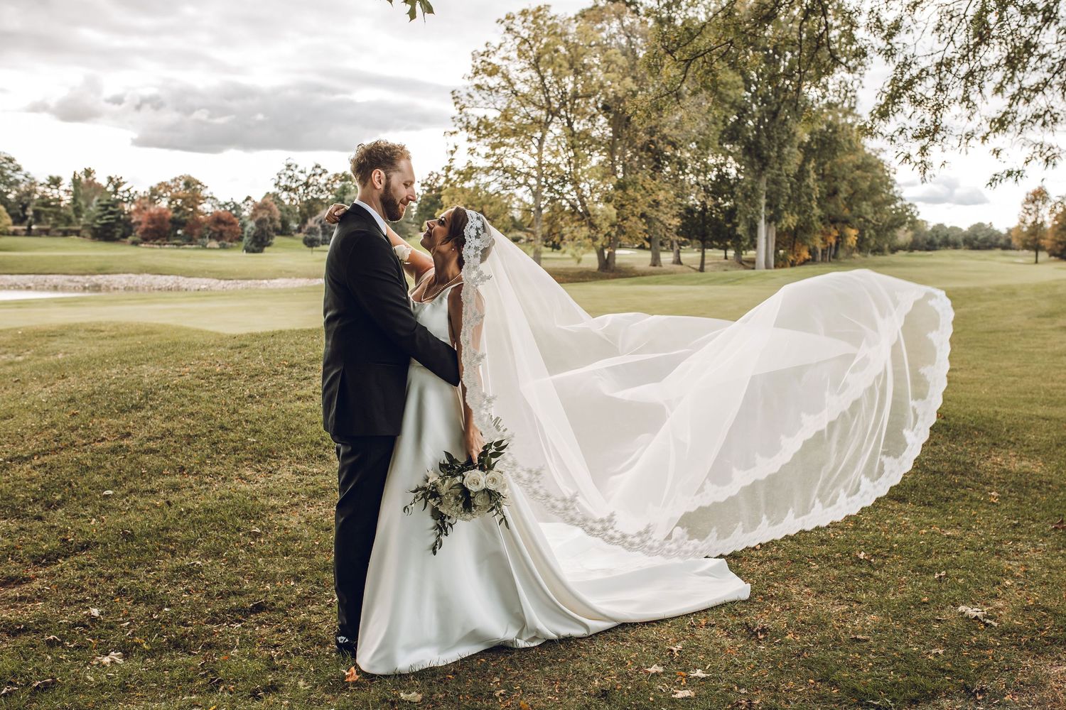 Bride and groom share romantic kiss outdoors, her veil flowing in wind, surrounded by lush green trees at golf course wedding.