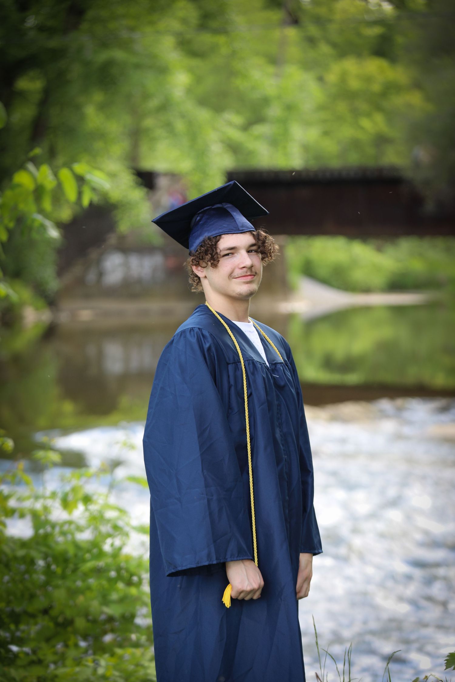 Young man in navy blue graduation cap and gown smiling outdoors near a creek with lush green trees.
