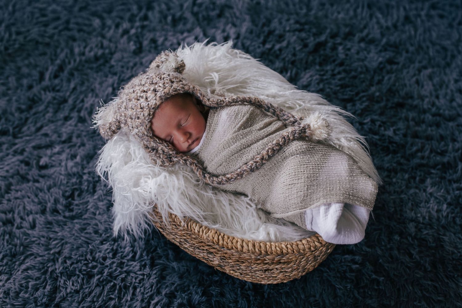Peaceful newborn baby sleeping in a wicker basket with fluffy white fur, wrapped in a knit swaddle and bear bonnet.