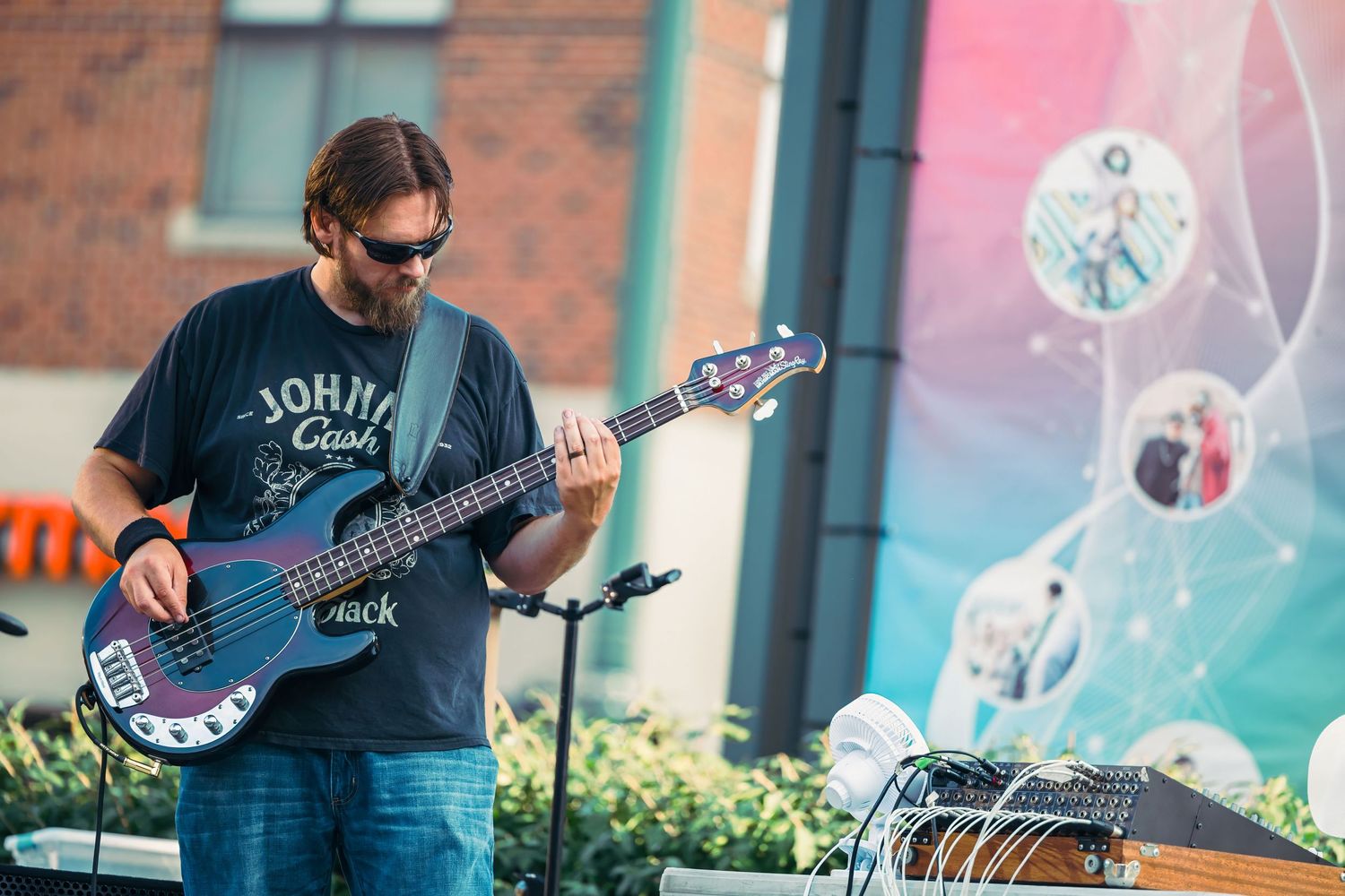 Musician playing electric bass guitar on outdoor stage wearing Johnny Cash t-shirt at a live music event.