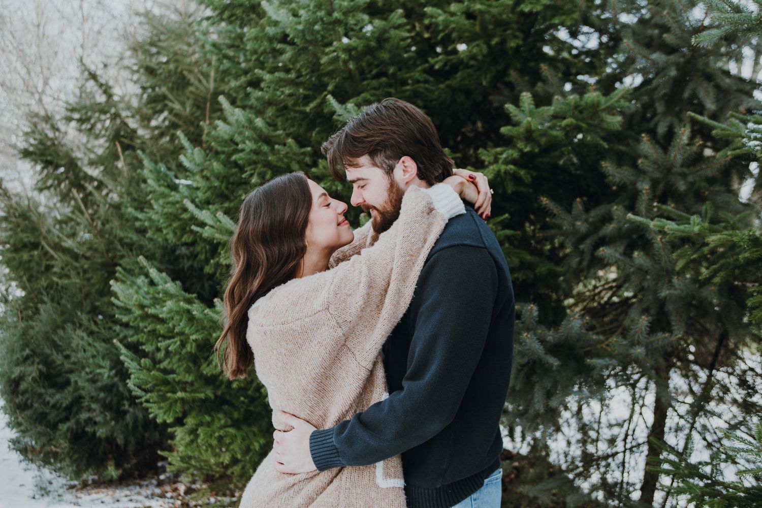 A winter embrace between two people wearing cozy sweaters among evergreen trees.