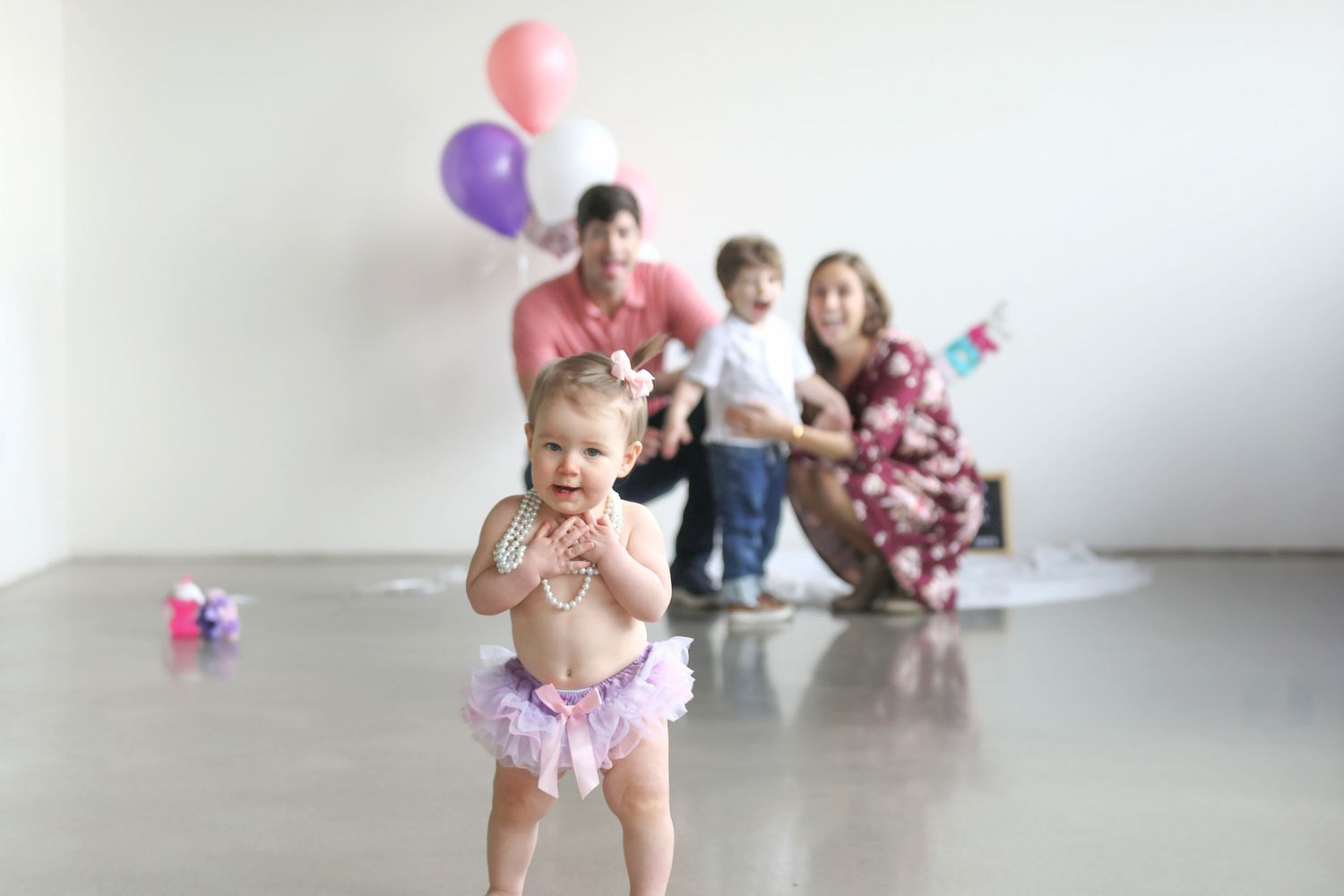 Baby in pink tutu stands at first birthday party with family and colorful balloons in bright white studio.