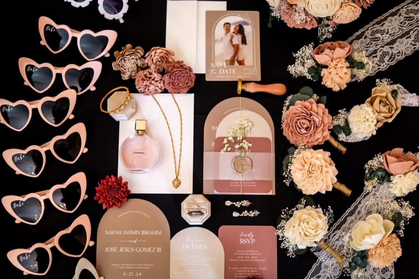 Wedding flat lay featuring heart sunglasses, floral bouquets, gold jewelry, invitation suite, and pink perfume on black background.