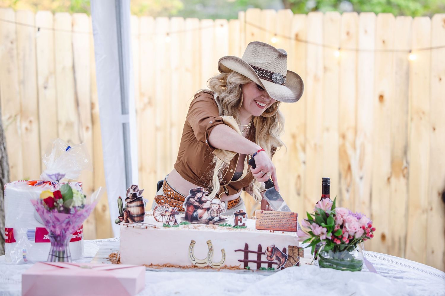 Woman in cowboy hat cutting a Western-themed cake at an outdoor party with floral decorations and wooden fence backdrop.