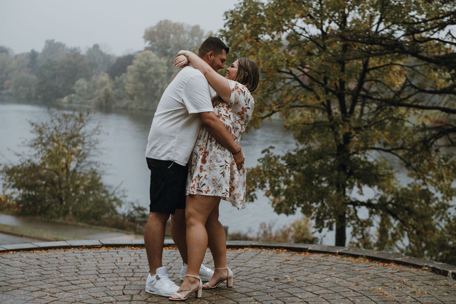 Couple embracing and dancing outdoors near a misty lake surrounded by autumn trees on a cobblestone overlook.
