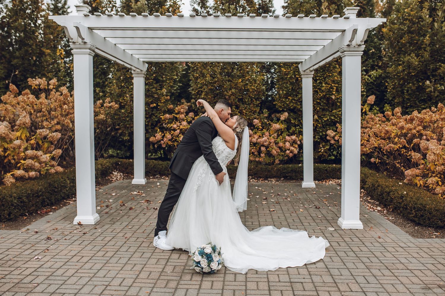 Bride and groom share a romantic dip kiss under a white pergola surrounded by autumn foliage in a garden wedding.