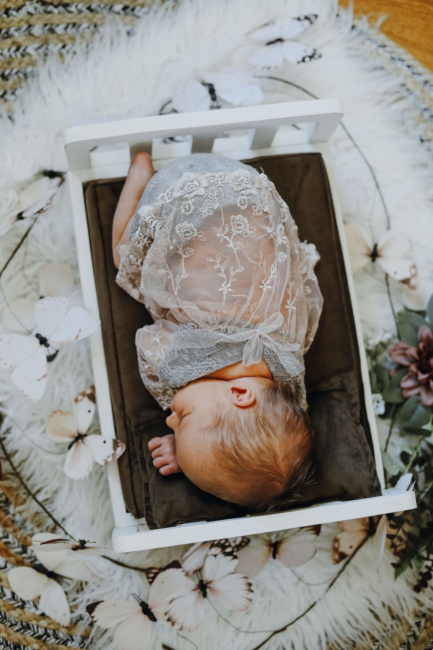 A small baby rests in a white wooden crate wearing a delicate vintage lace bonnet on a floral background.