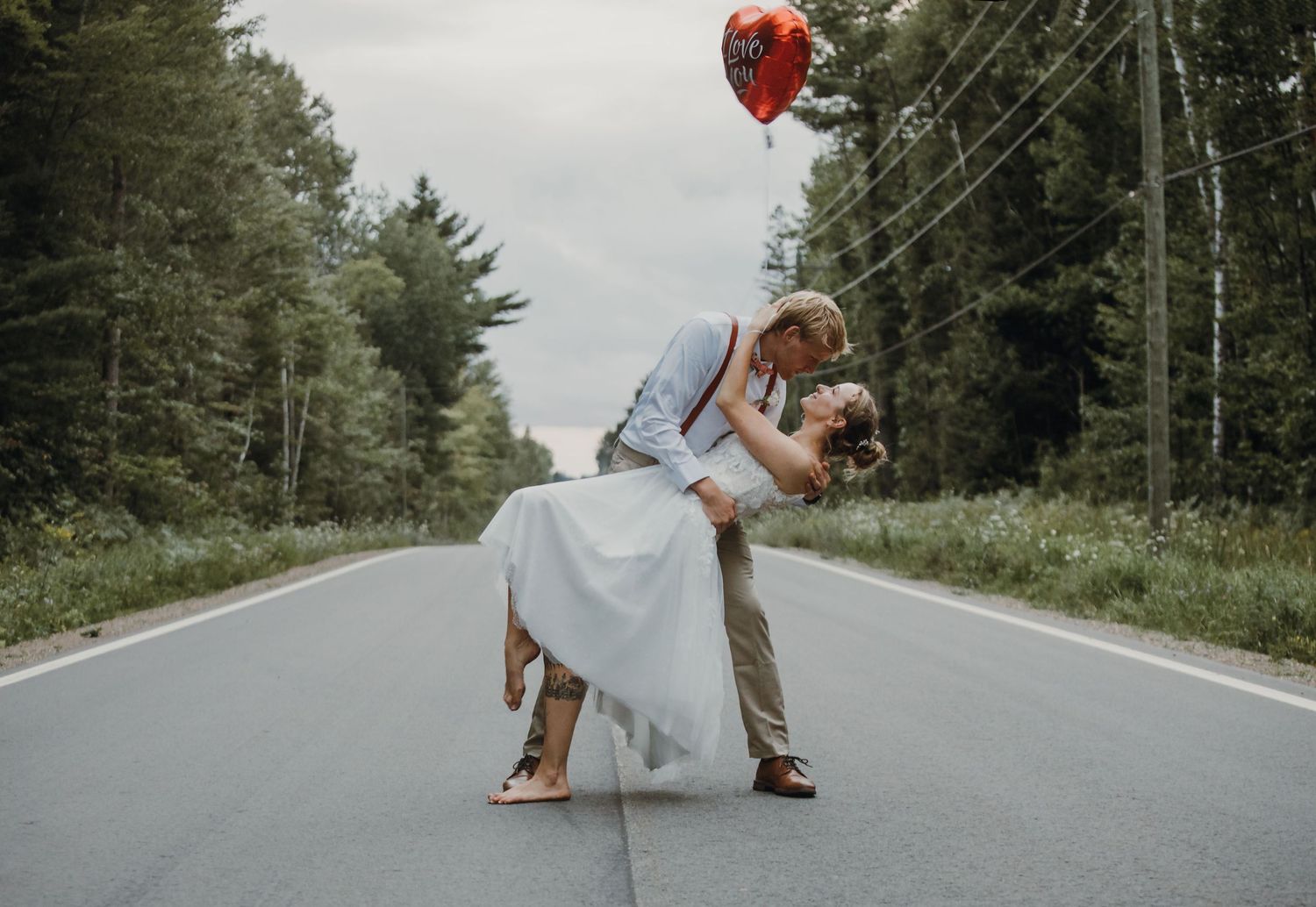 A couple in wedding attire dances and twirls on an empty forest road surrounded by evergreen trees.