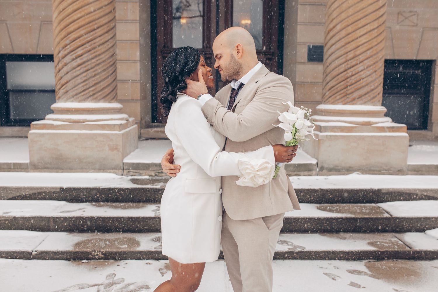 A couple in matching white and beige outfits embrace on snowy steps outside a building with ornate columns.