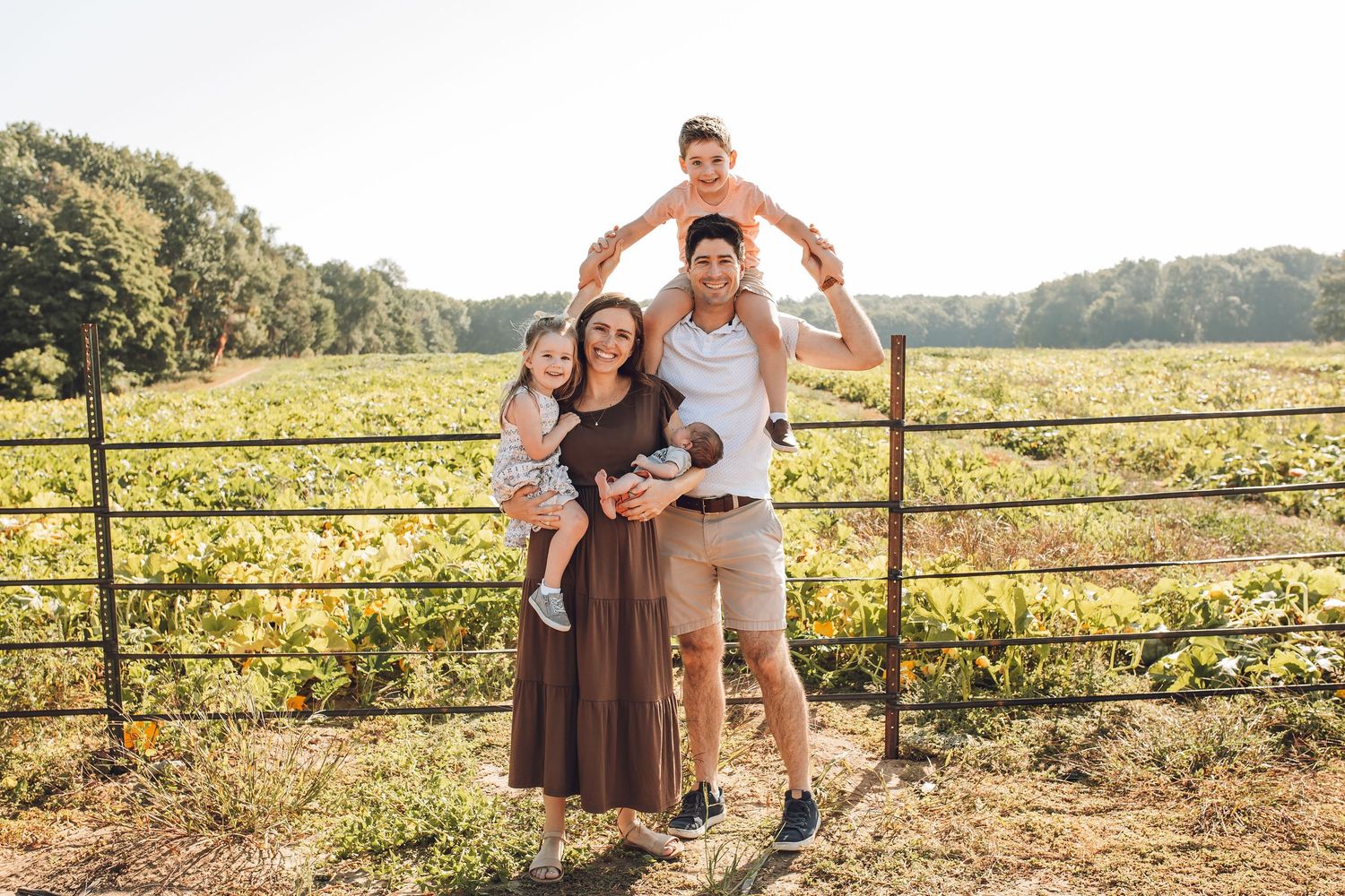 Family of four posing by a farm fence in a sunny field, parents holding young children with green crops in background.