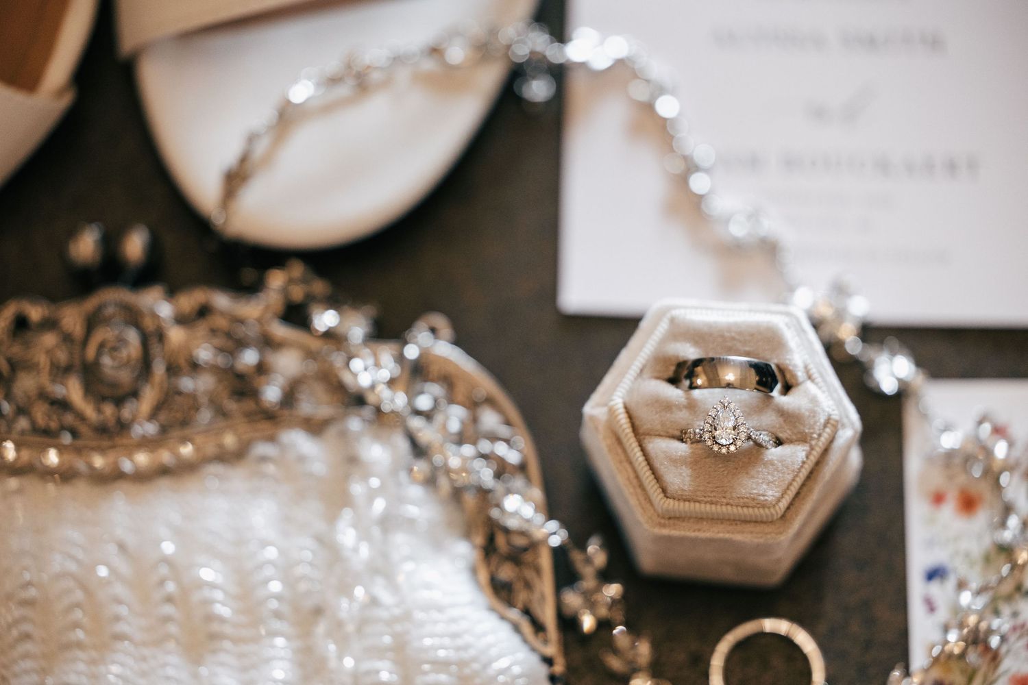 Elegant wedding flat lay featuring diamond engagement ring in velvet box, bridal accessories, tiara, and invitation on dark background.