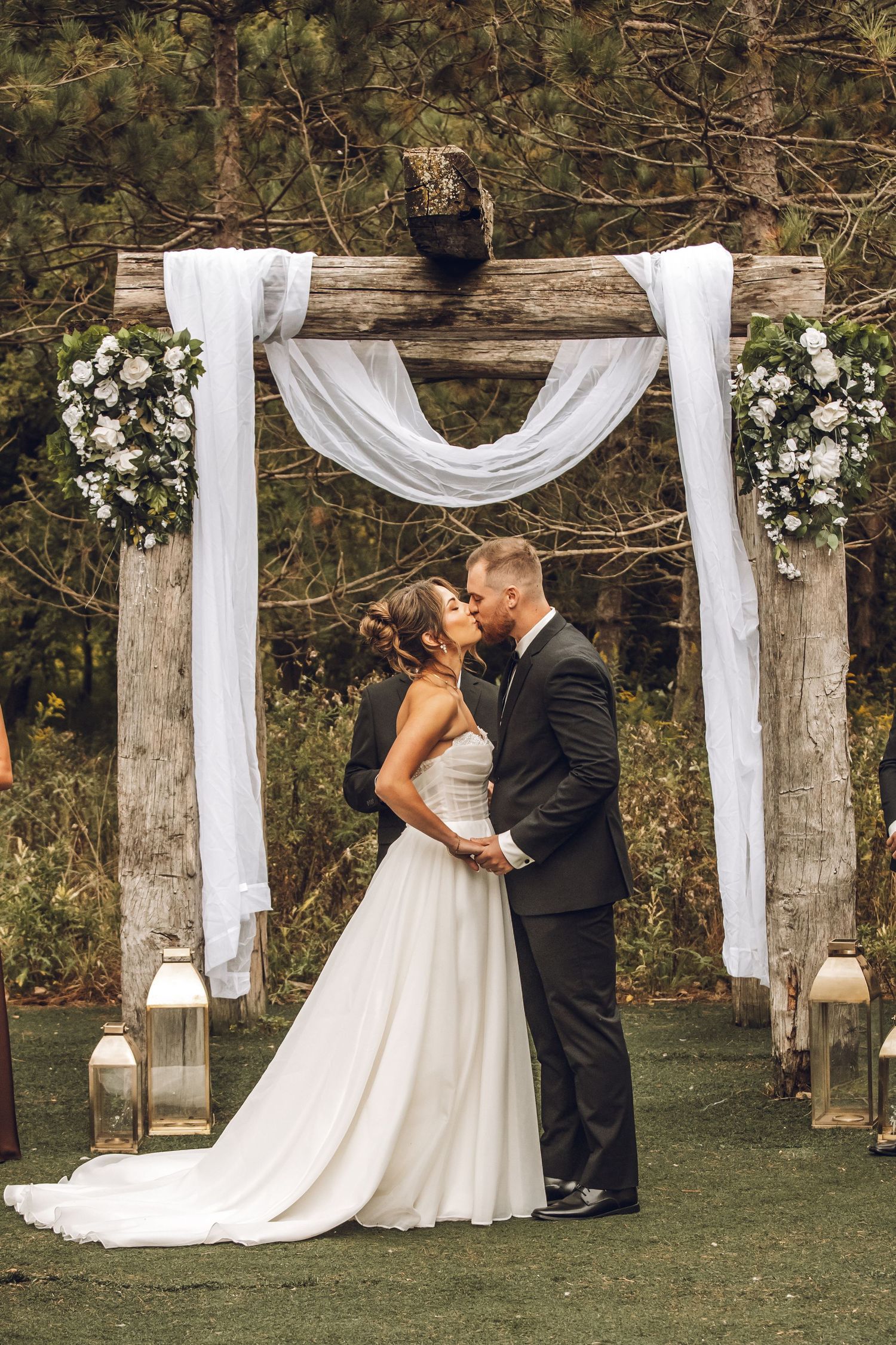 A romantic outdoor wedding ceremony with wooden posts draped in white fabric and floral arrangements in a wooded setting.