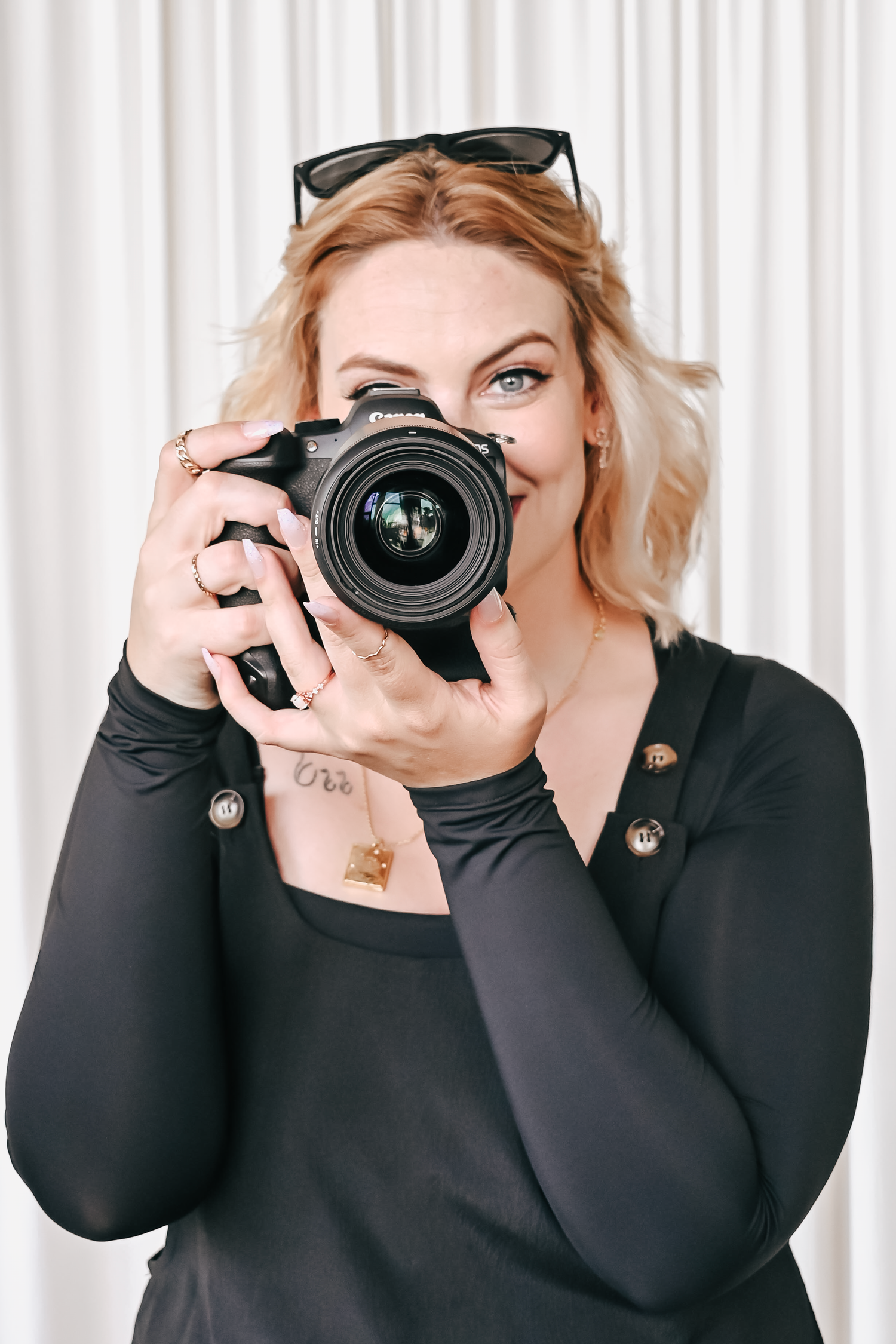 A photographer holding a professional camera with lens up to their face against white curtain background.