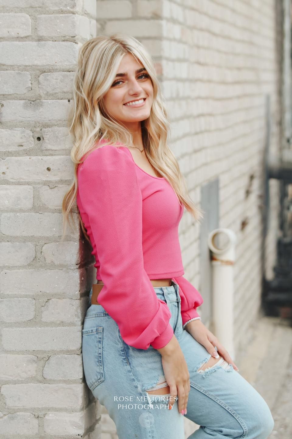 A smiling blonde woman in a pink crop top and ripped jeans leans against a white brick wall.