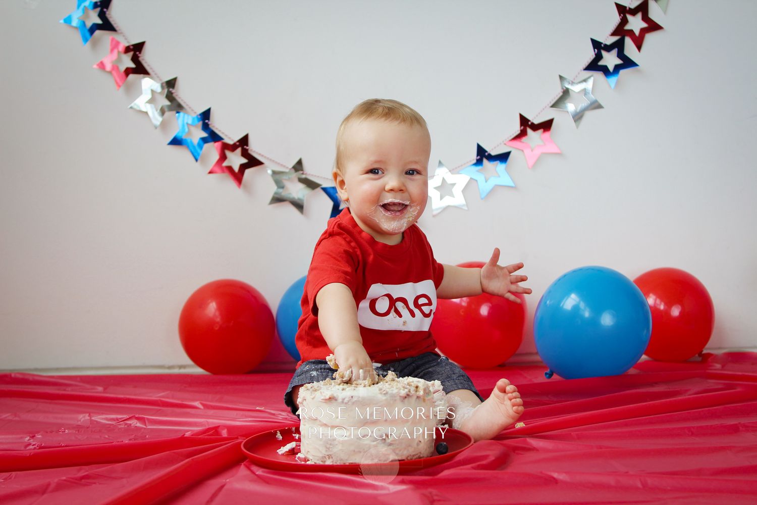 Baby boy in red shirt smashes first birthday cake surrounded by red and blue balloons and star garland.