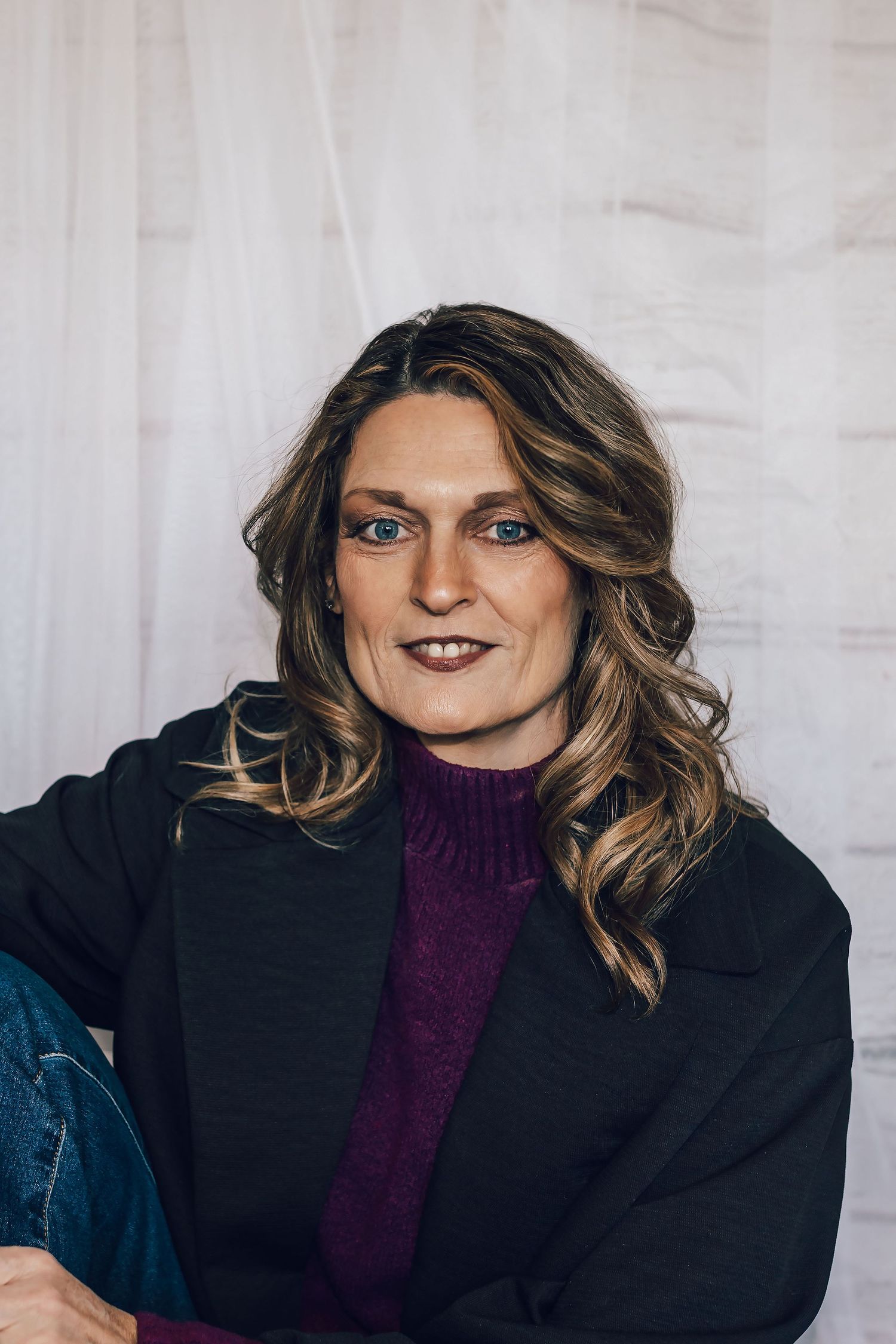 Woman with wavy highlighted hair wearing a purple turtleneck and black jacket, sitting casually against a white backdrop.