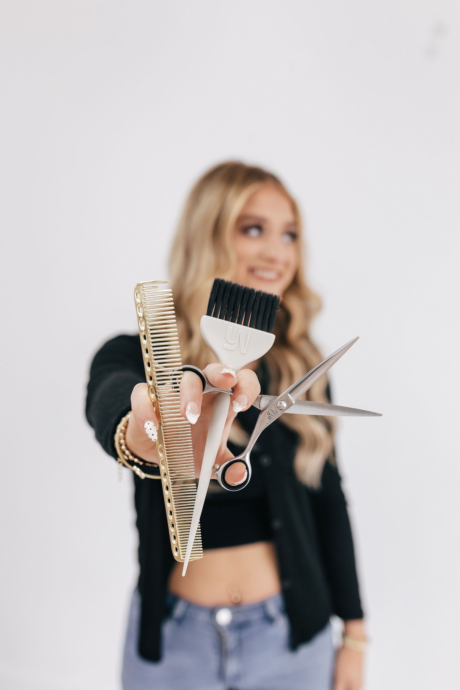 Blonde hairstylist holding gold comb, dye brush, and scissors toward camera against white background.