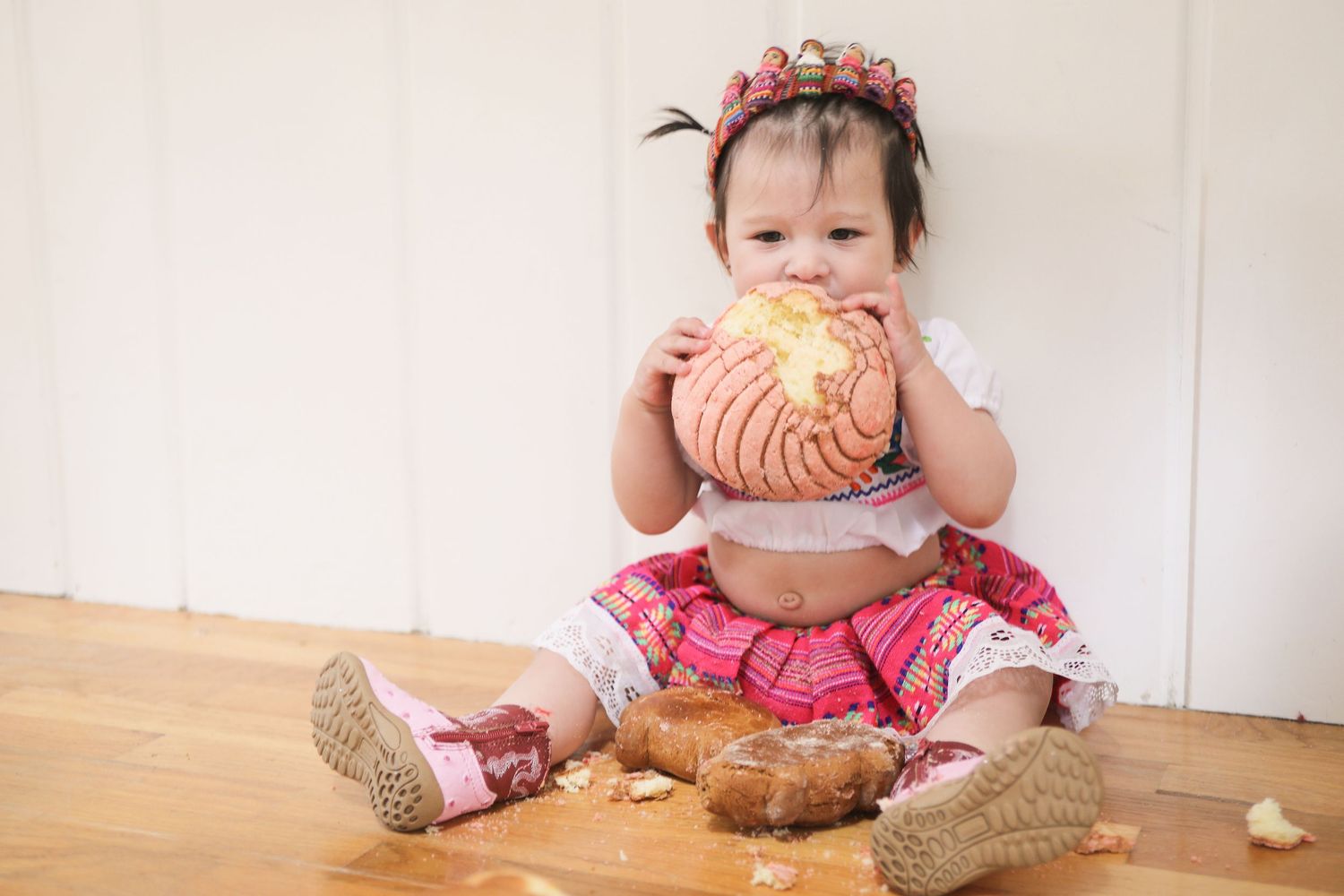 Adorable baby girl in traditional pink outfit sitting on floor eating a large bread roll during smash cake photoshoot.