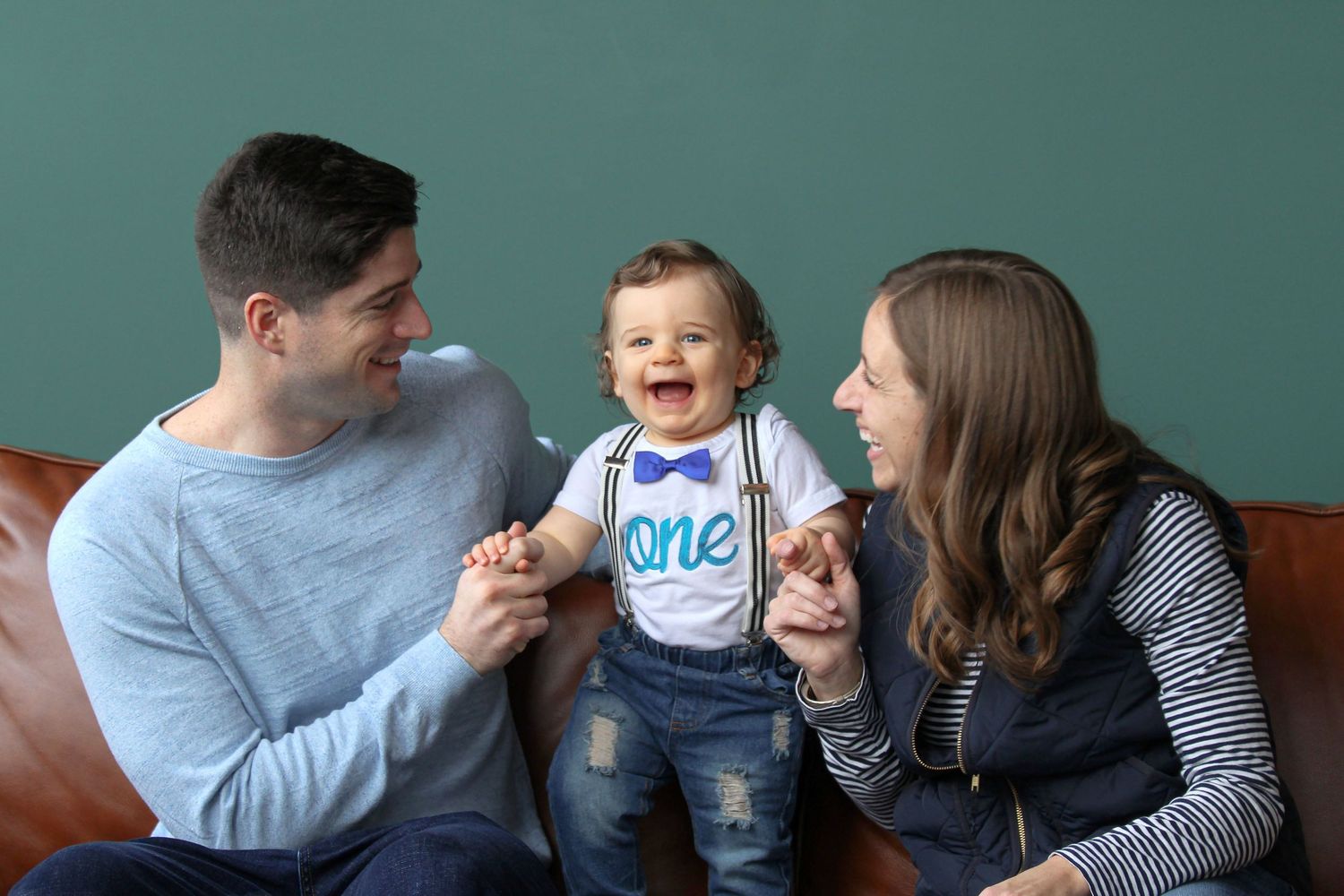 Happy family celebrating baby's first birthday, parents smiling at laughing toddler wearing 'one' outfit with bow tie.