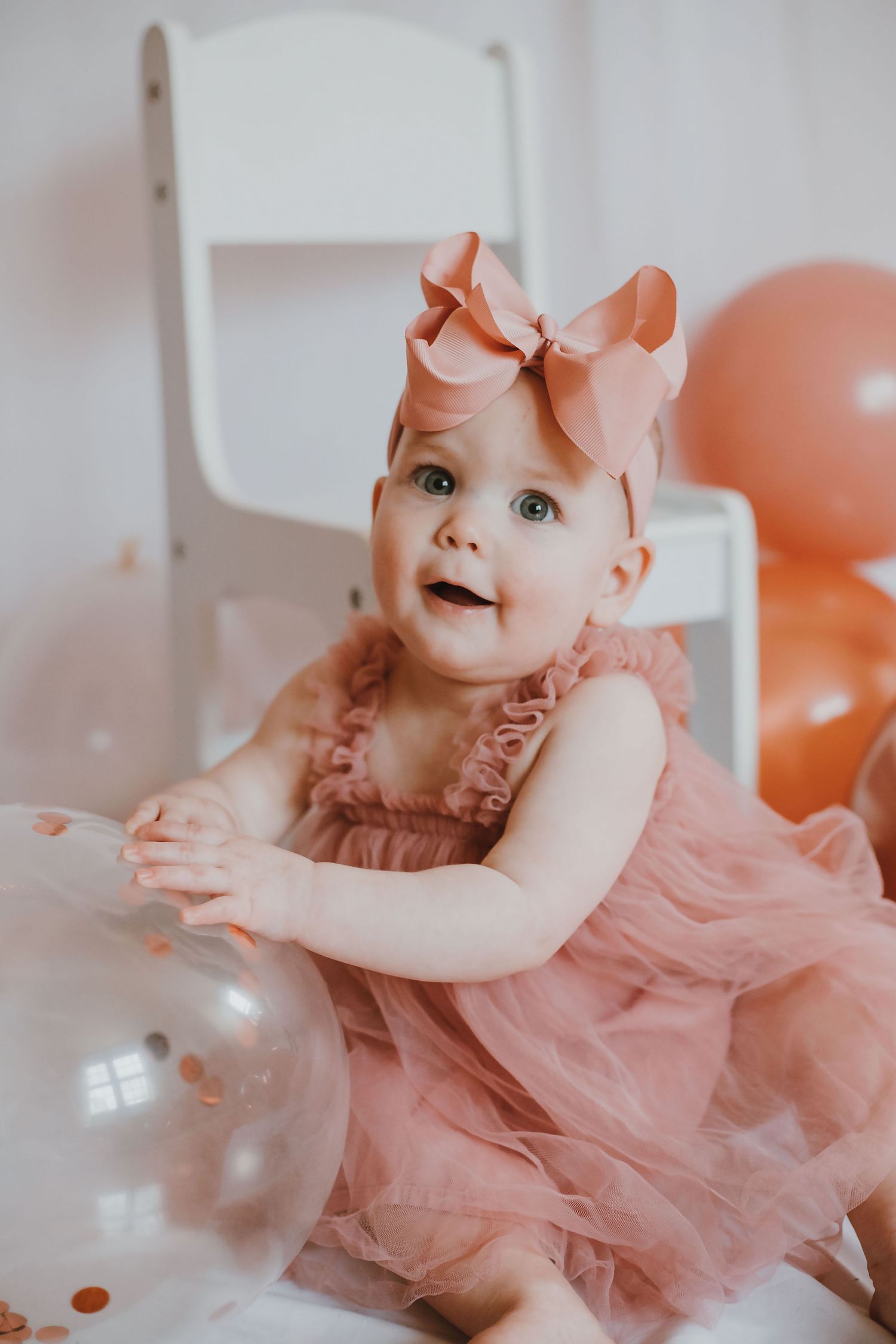Adorable baby girl in pink tulle dress and bow headband sitting with balloons at first birthday party.