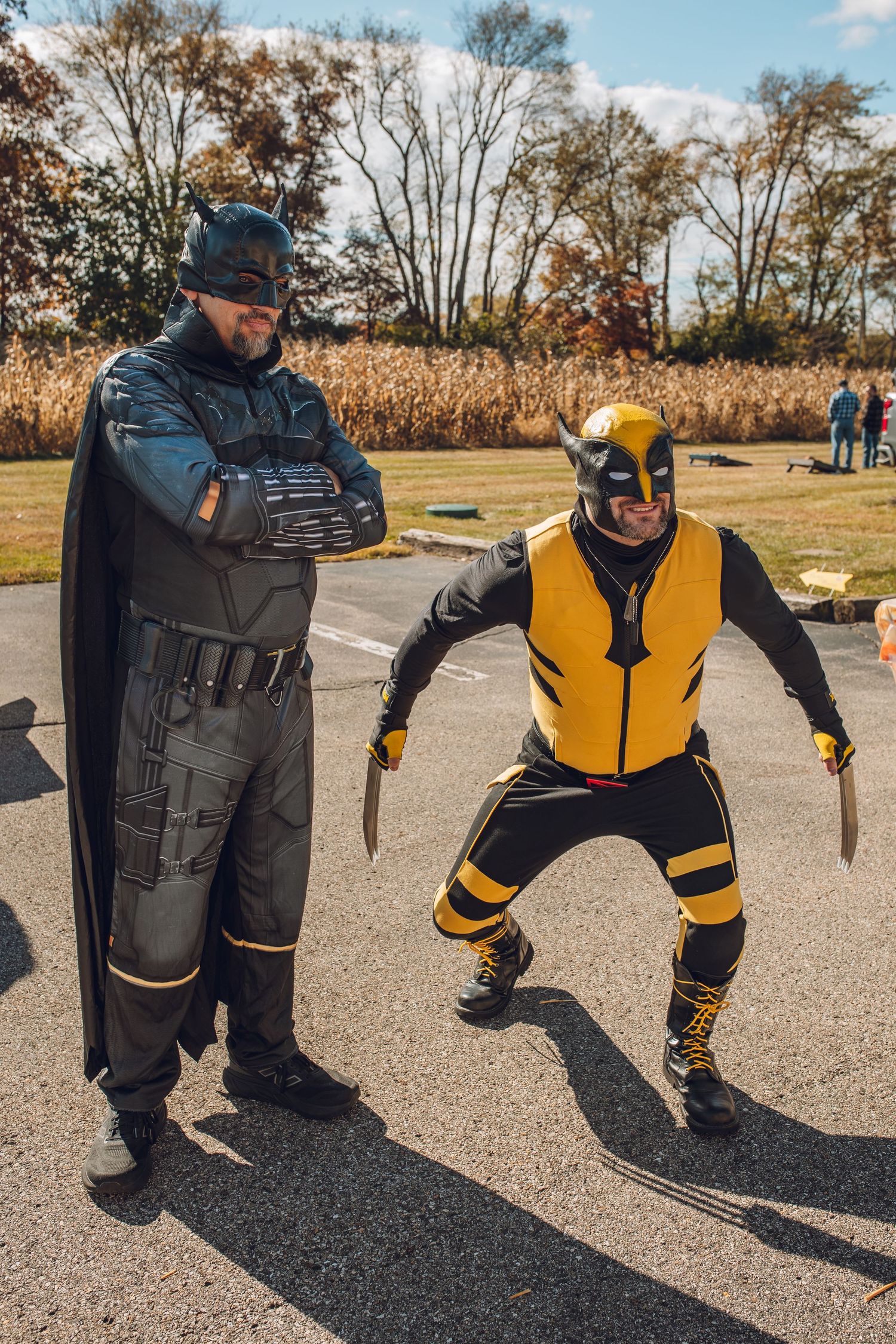 Two men in Batman and Wolverine costumes pose outdoors in a parking lot on a sunny autumn day.
