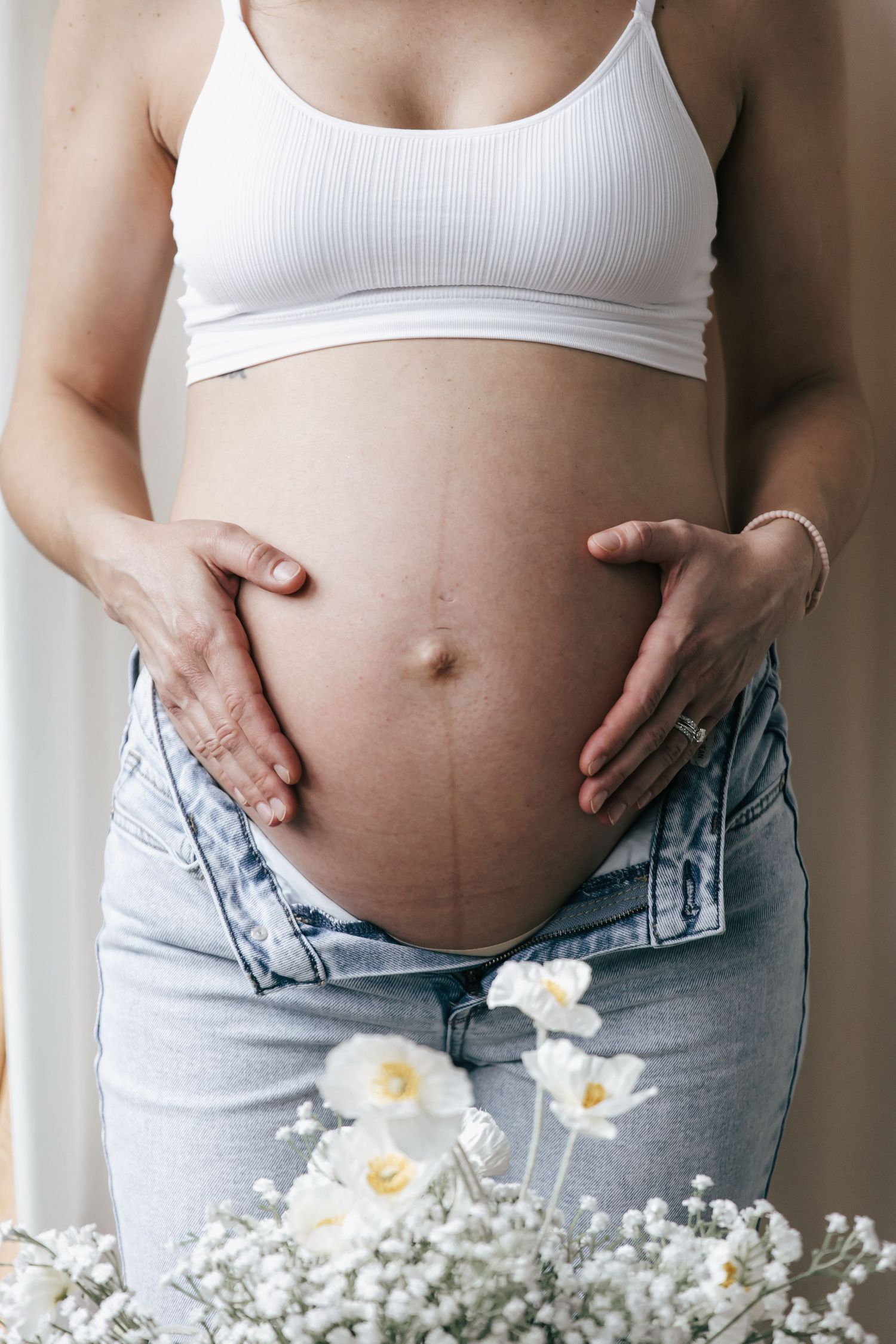 Pregnant woman in white top and jeans holding her belly with white flowers in foreground.