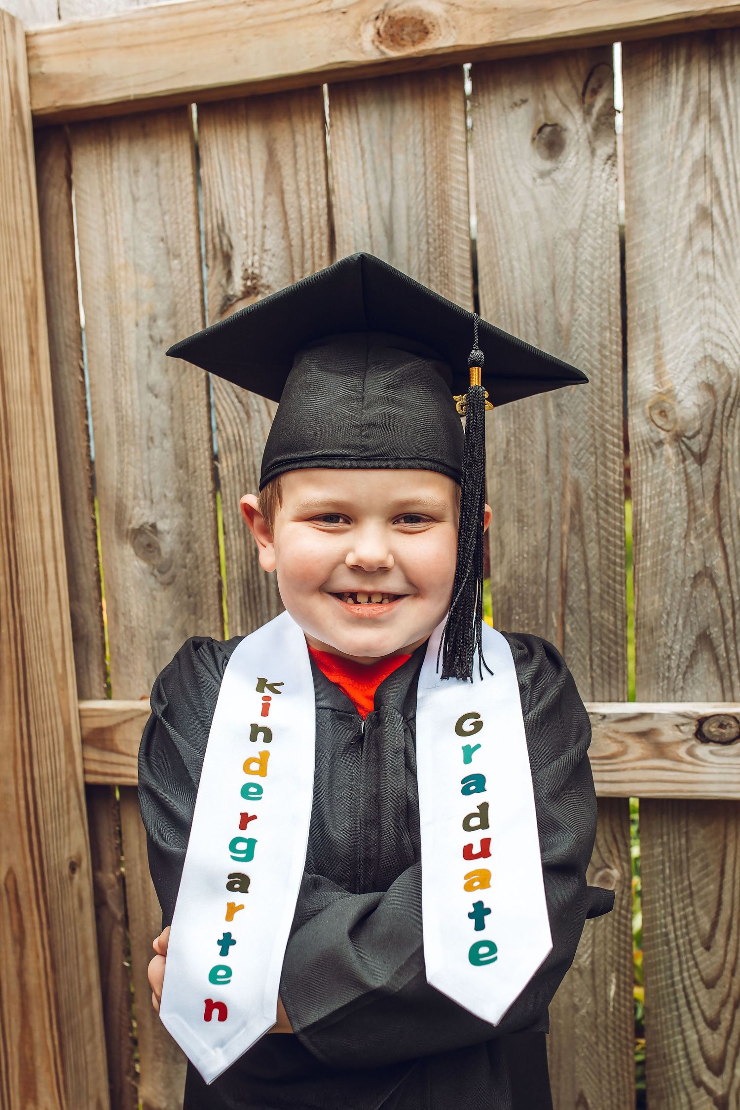 Smiling child in black graduation cap and gown wearing a Kindergarten Graduate stole, posing outdoors.
