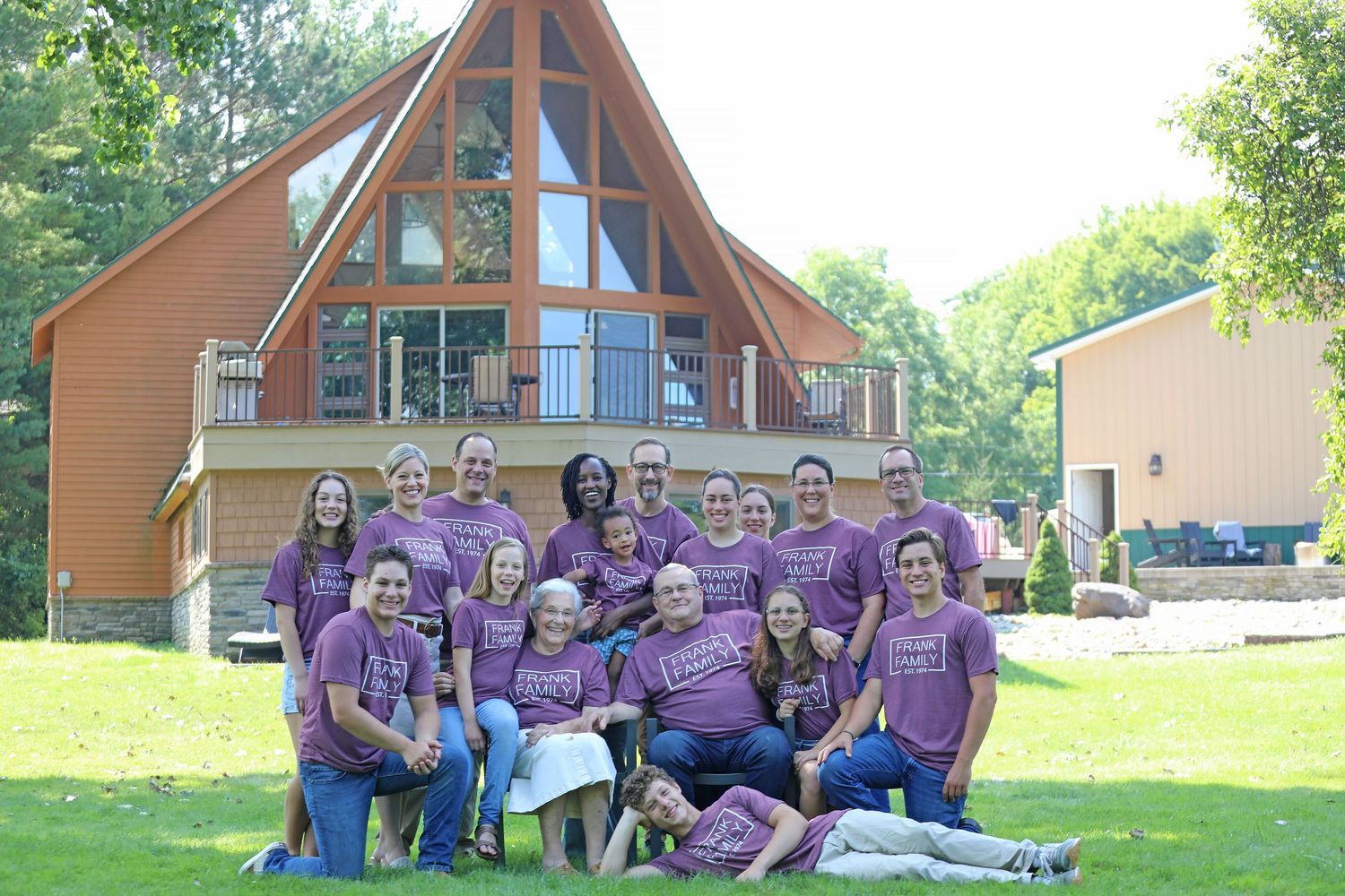Large family group in matching purple t-shirts posing outdoors in front of a wooden A-frame cabin surrounded by trees.