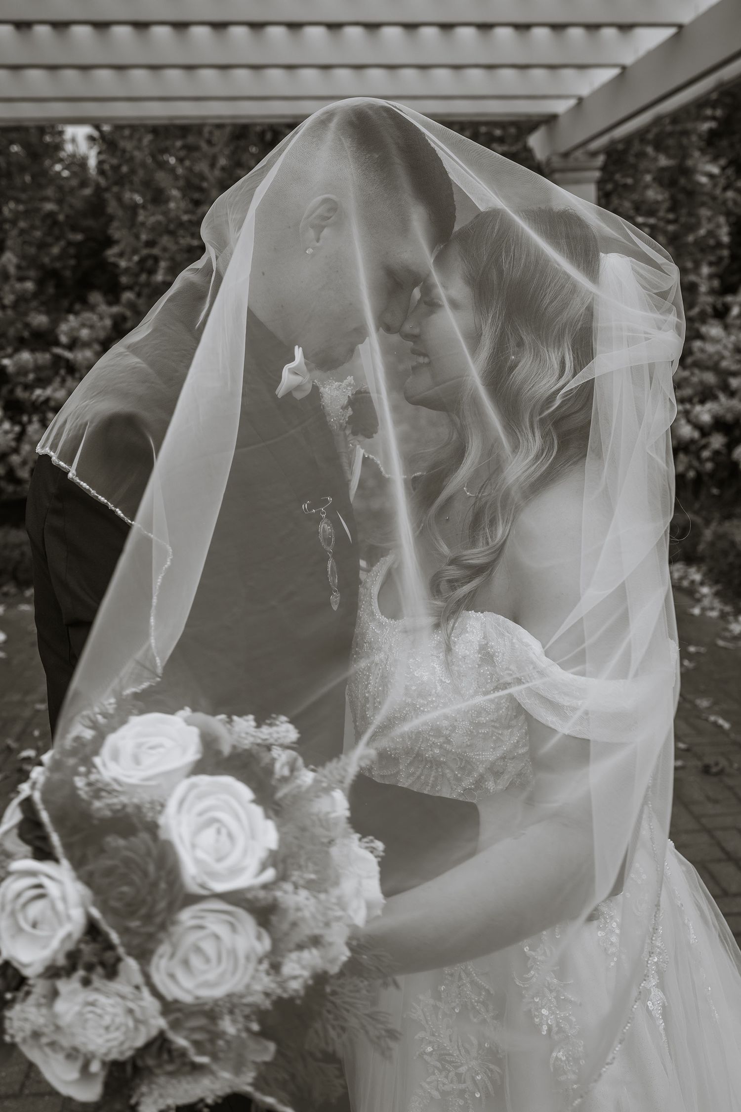 Black and white photo of bride and groom kissing under wedding veil, bride holding rose bouquet.