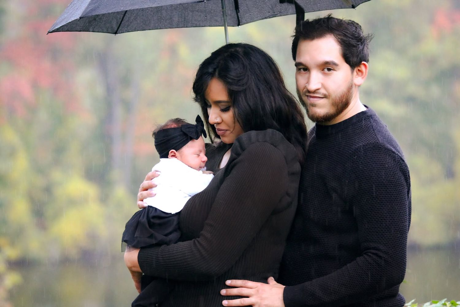 A couple holding a newborn baby under an umbrella in a rainy autumn park setting.