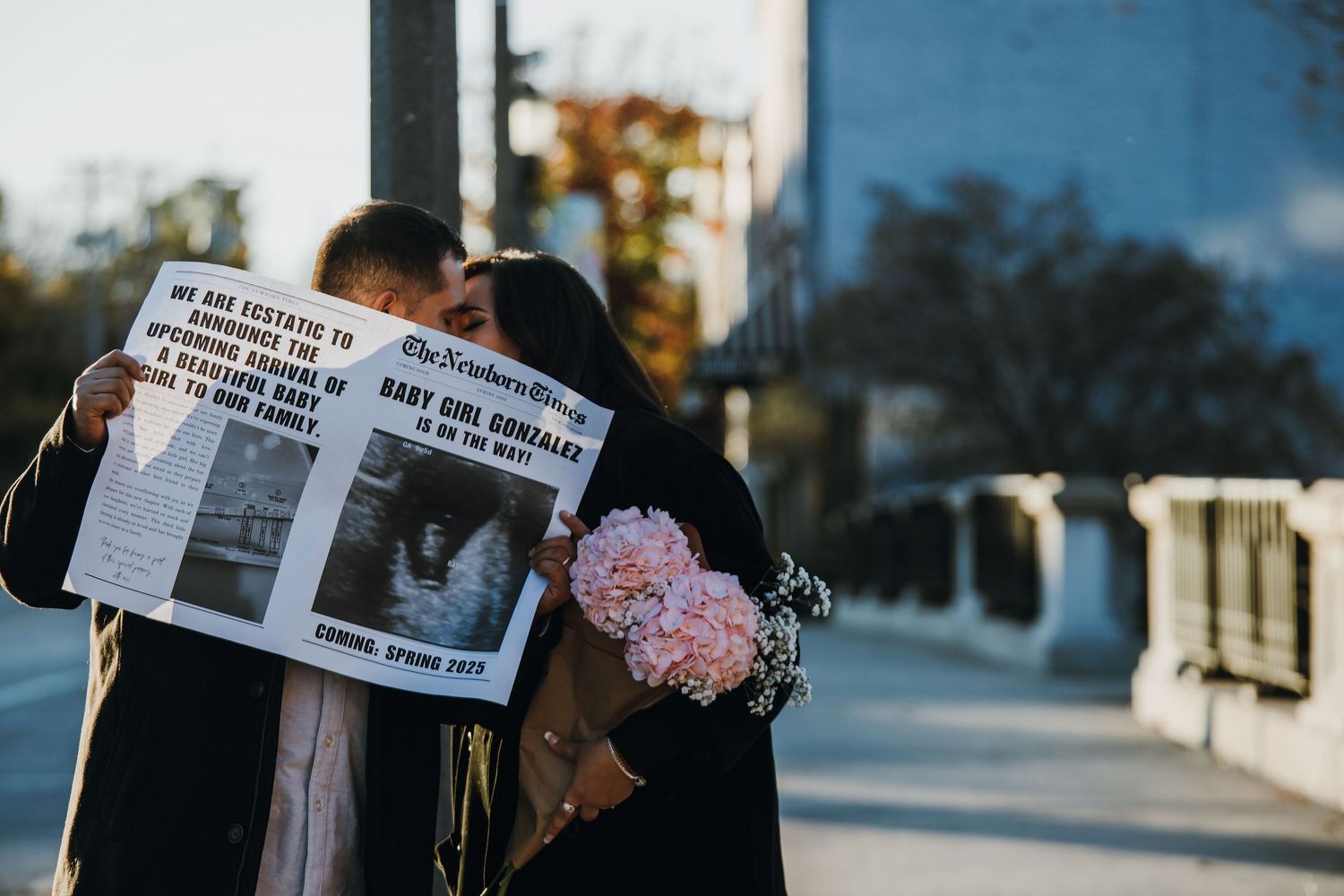 Couple embracing while reading a custom pregnancy announcement newspaper, woman holding pink hydrangea bouquet outdoors.