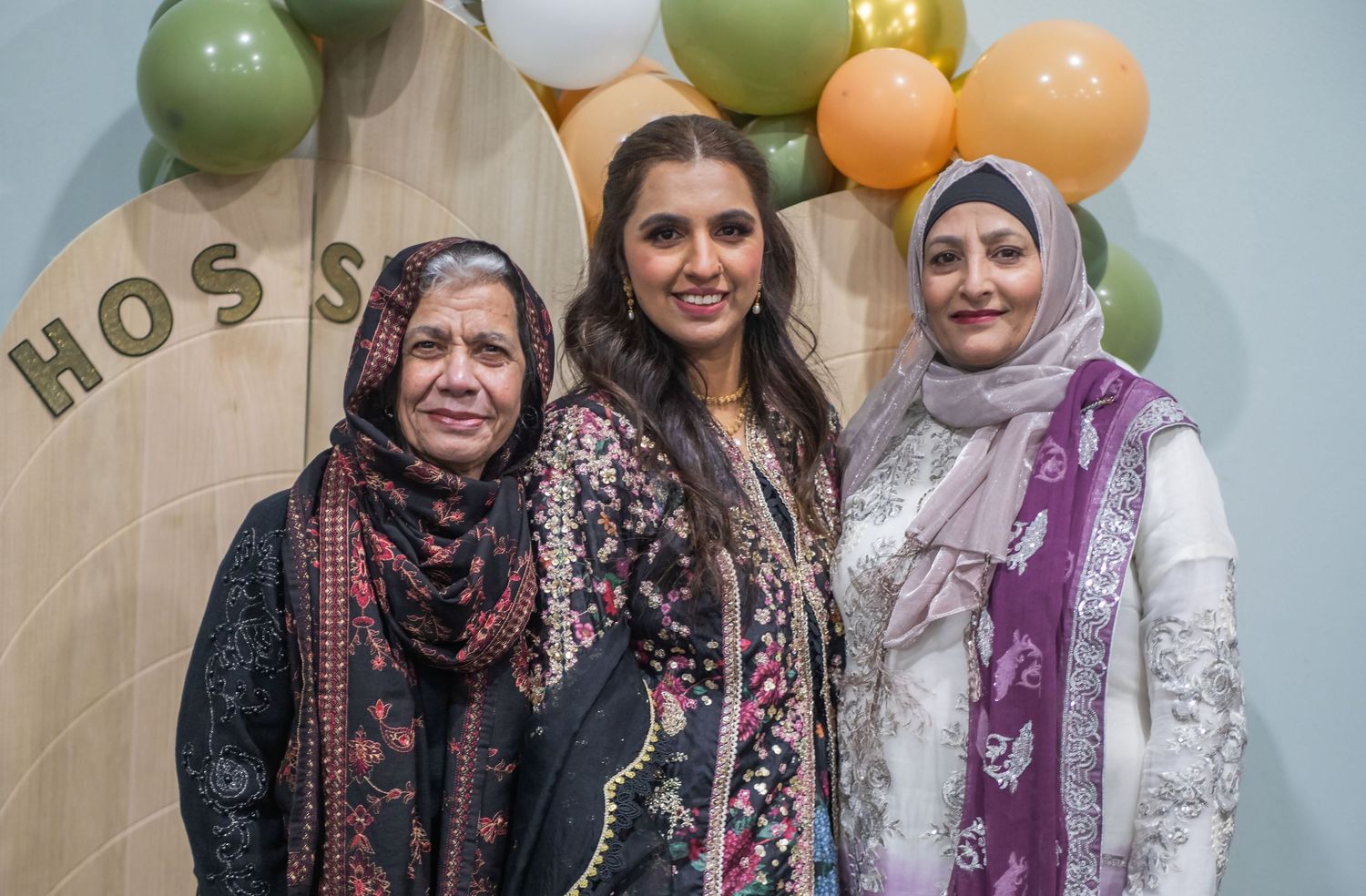 Three women in traditional South Asian attire smile together at a festive event with colorful balloons.