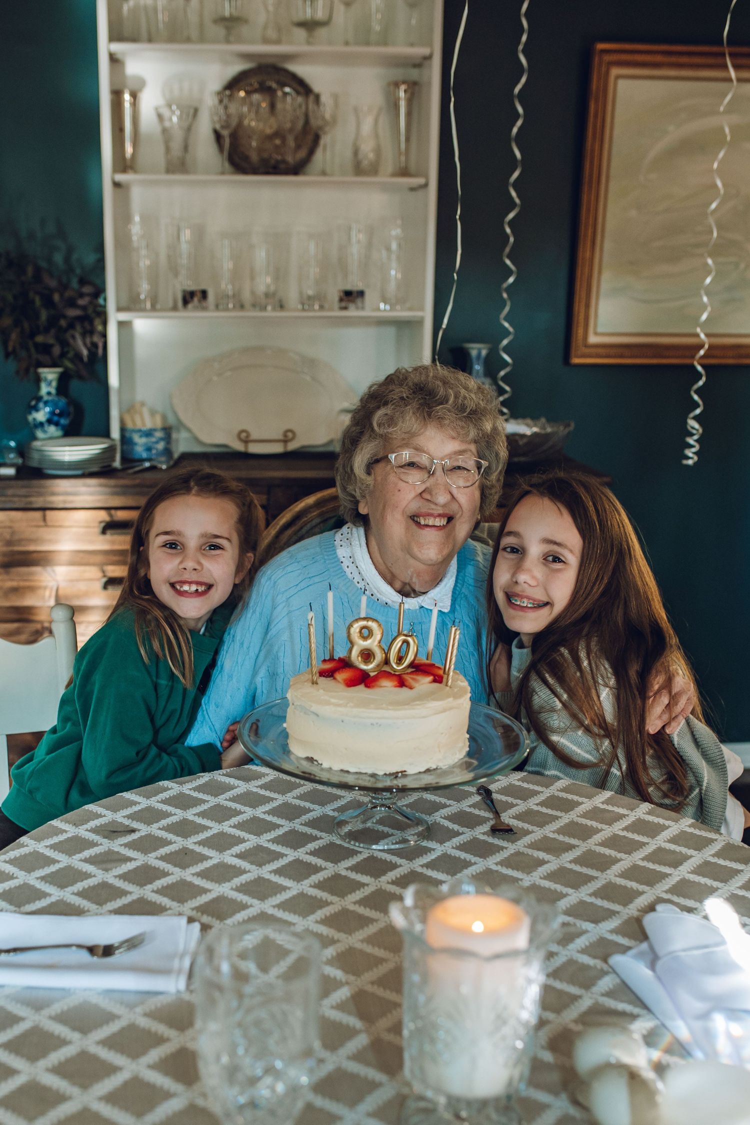 Elderly woman celebrating her 80th birthday with two granddaughters beside a white cake with candles at a family dinner.