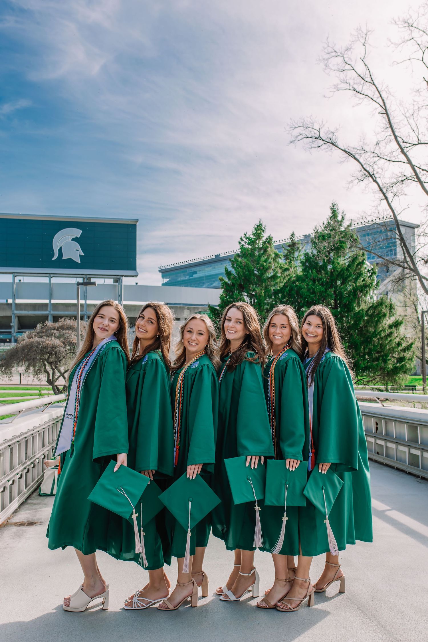 Six Michigan State University graduates pose in green caps and gowns on campus with Spartan logo visible.