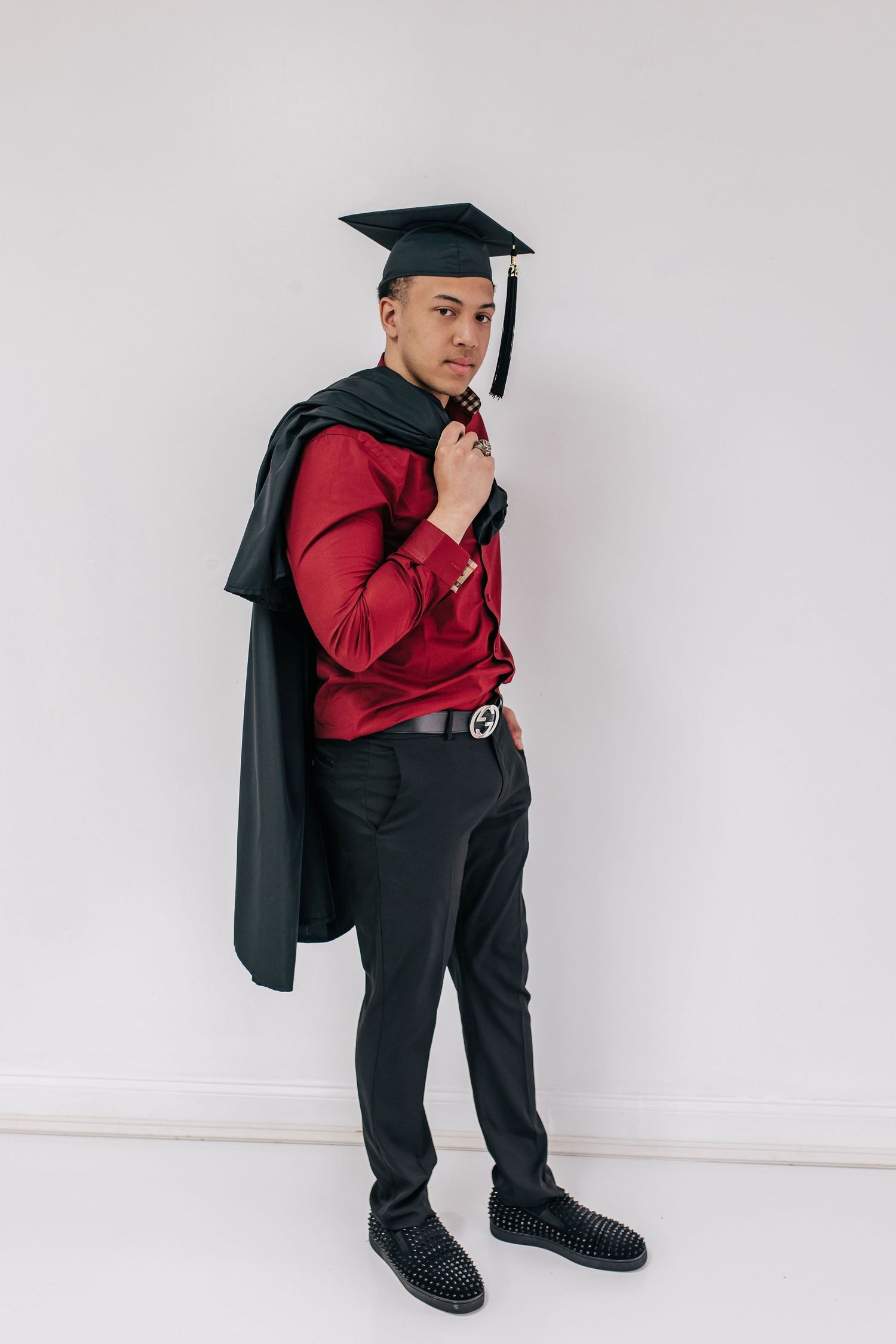 Young man in graduation cap and gown over red shirt and black pants posing against white wall.
