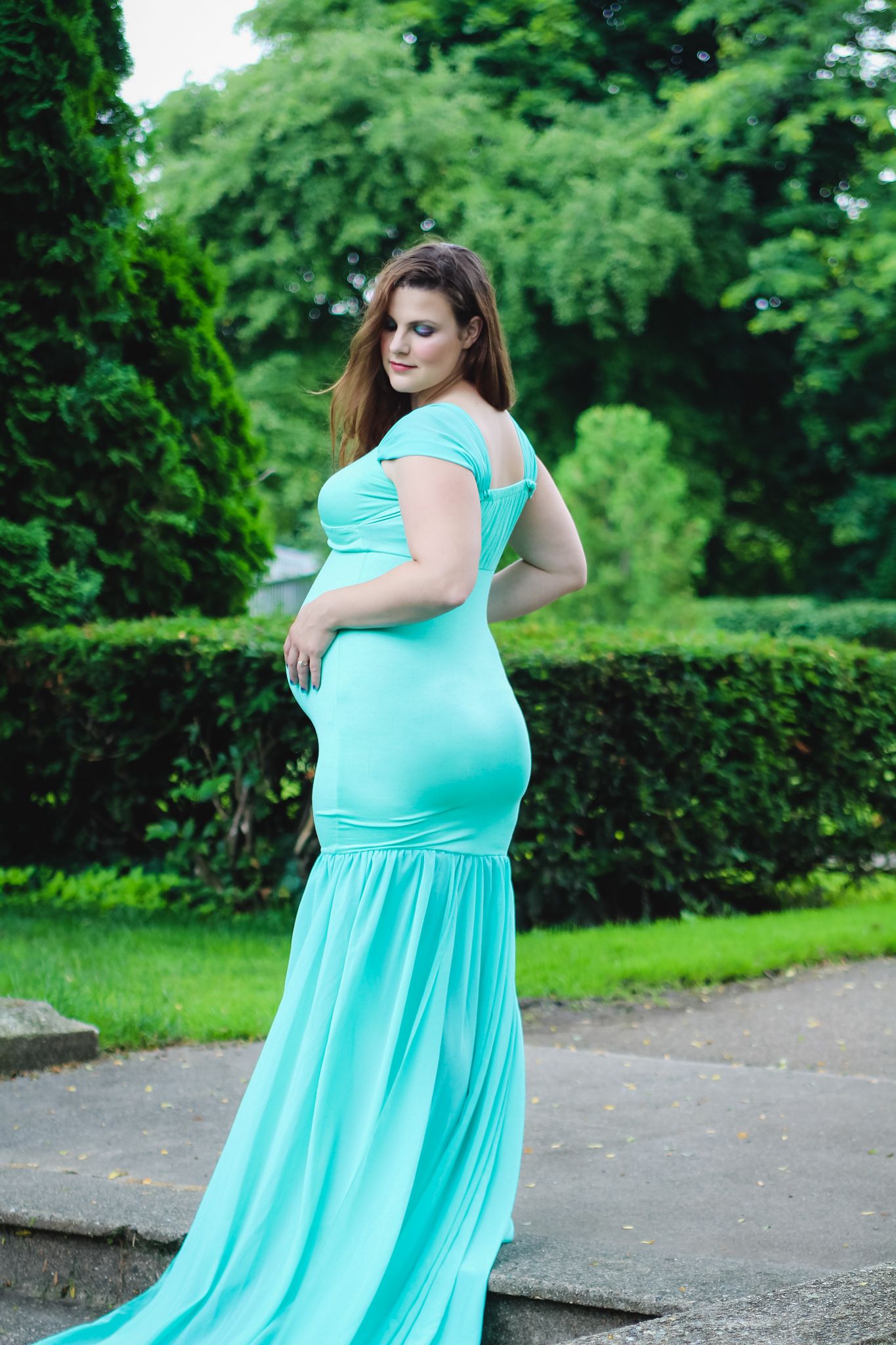 Pregnant woman in elegant teal mermaid gown posing gracefully in a lush green garden park setting.