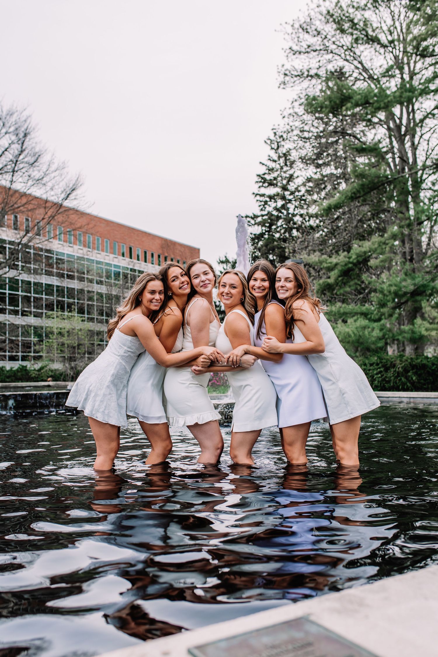 Seven young women in white dresses pose together smiling in a shallow reflecting pool on a college campus.