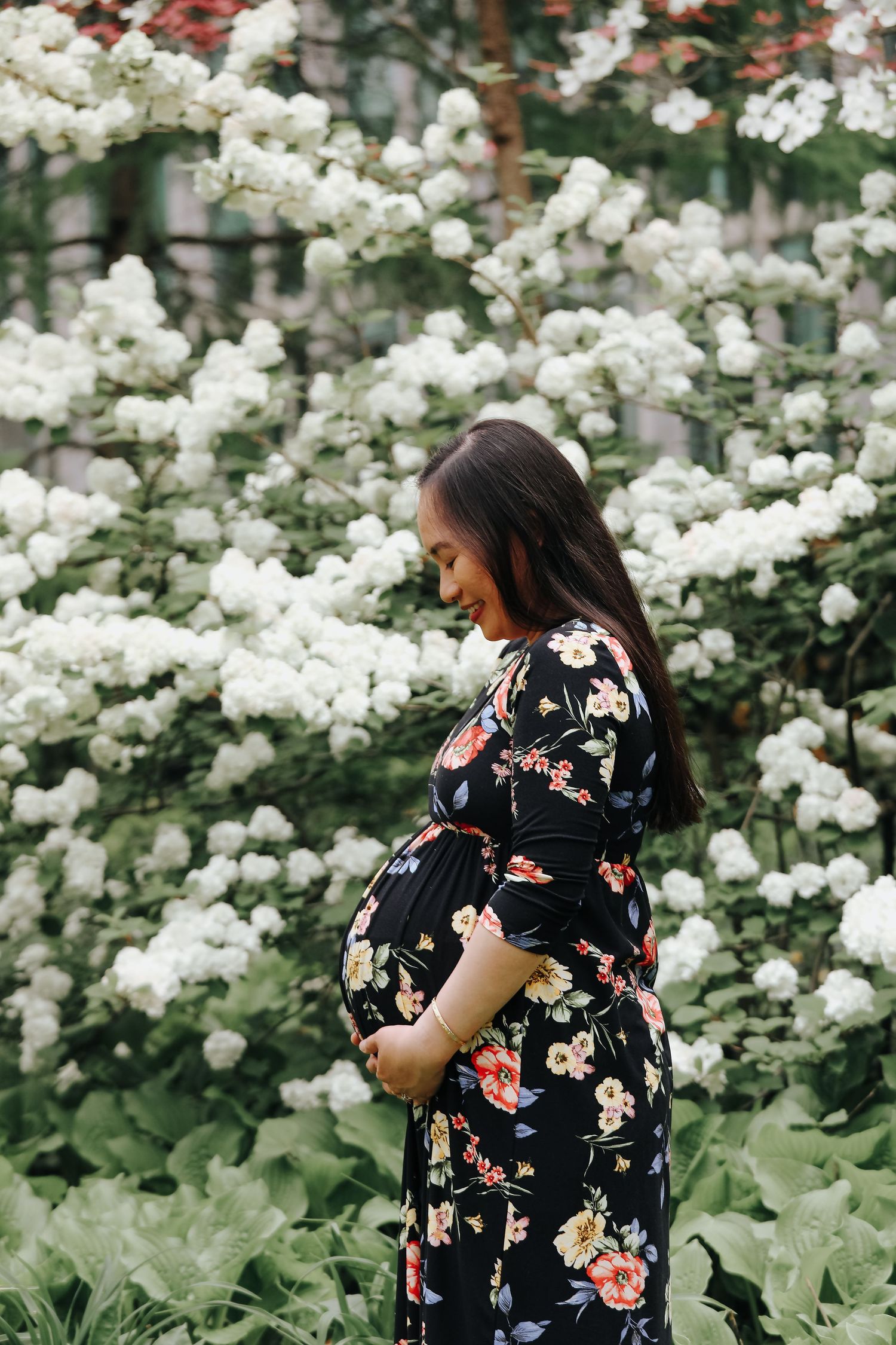 Pregnant woman in floral dress cradling baby bump, smiling softly among white blooming flowers outdoors.