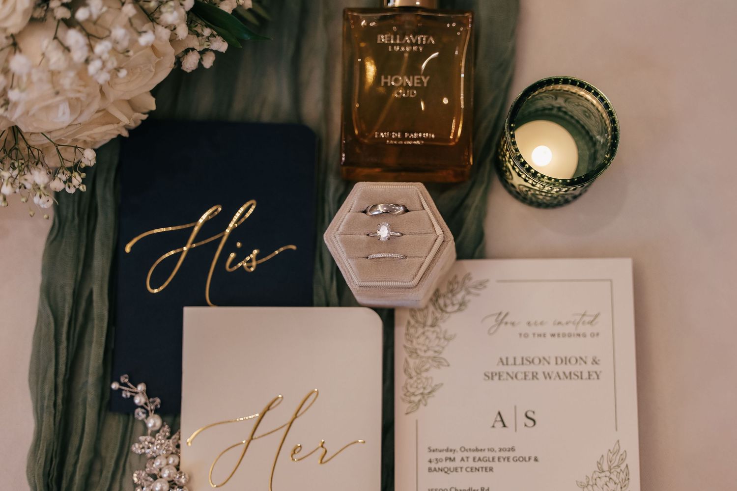 Elegant wedding flat lay featuring navy vow books, diamond ring, gold perfume, green candle, and floral invitation suite.