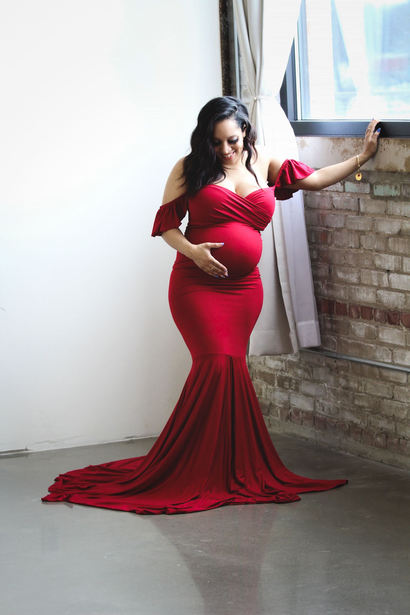 Pregnant woman in elegant red mermaid gown posing near brick wall window during maternity photo shoot.