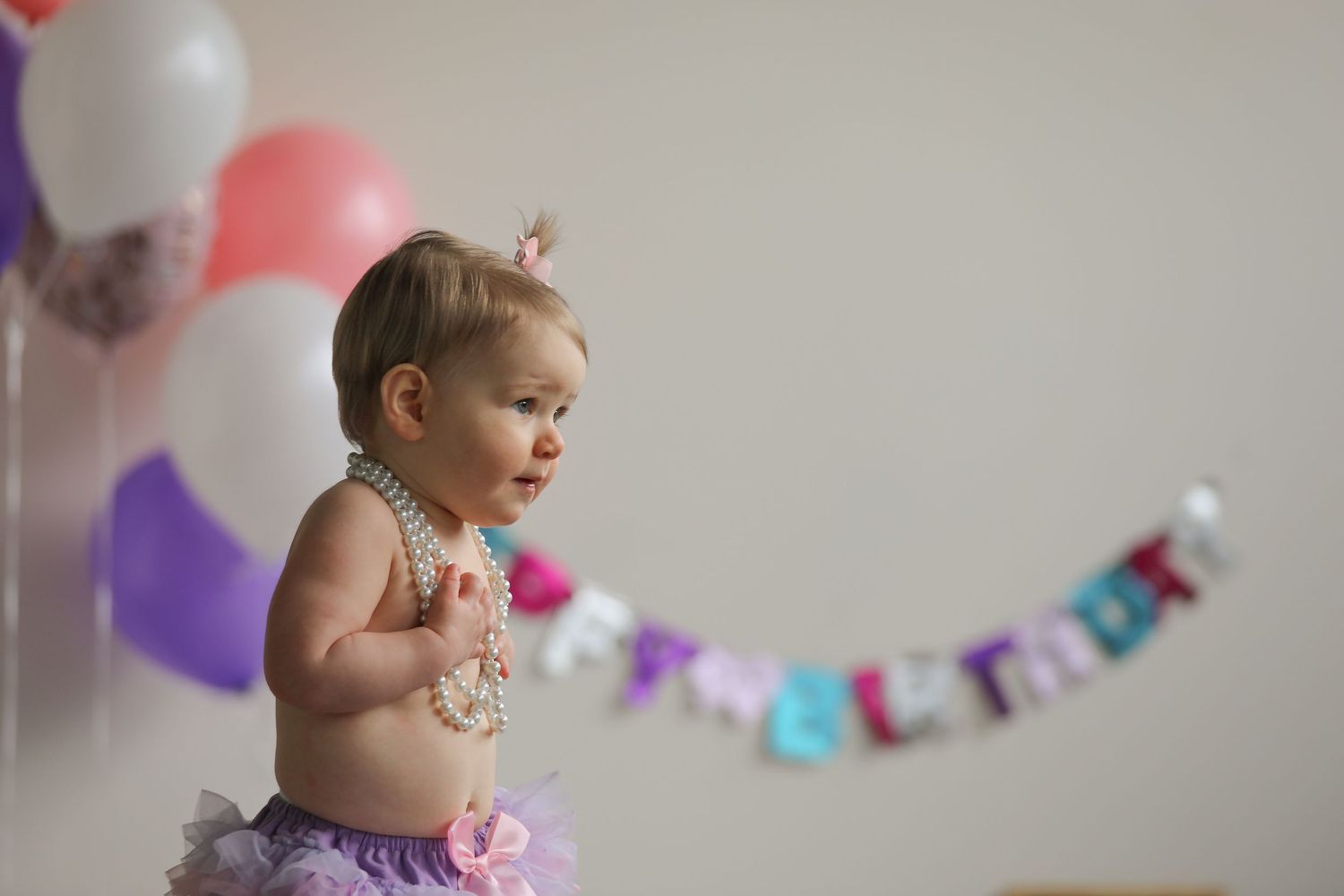 Baby girl in tutu skirt and pearl necklace celebrates first birthday with colorful balloons and banner.