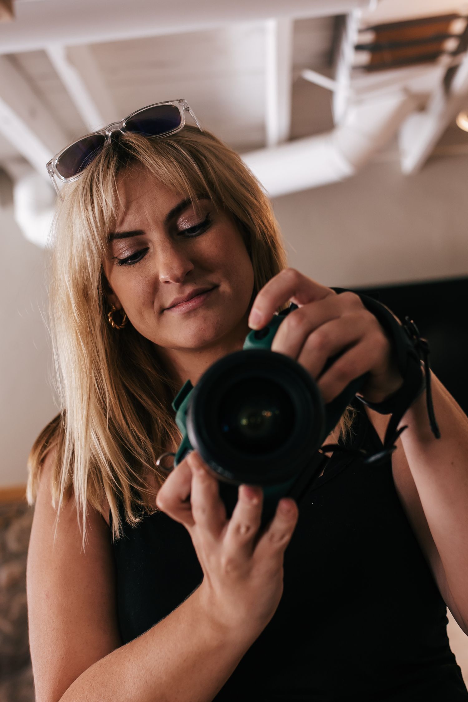 Blonde woman with sunglasses on head reviewing photos on DSLR camera, wearing black outfit indoors.