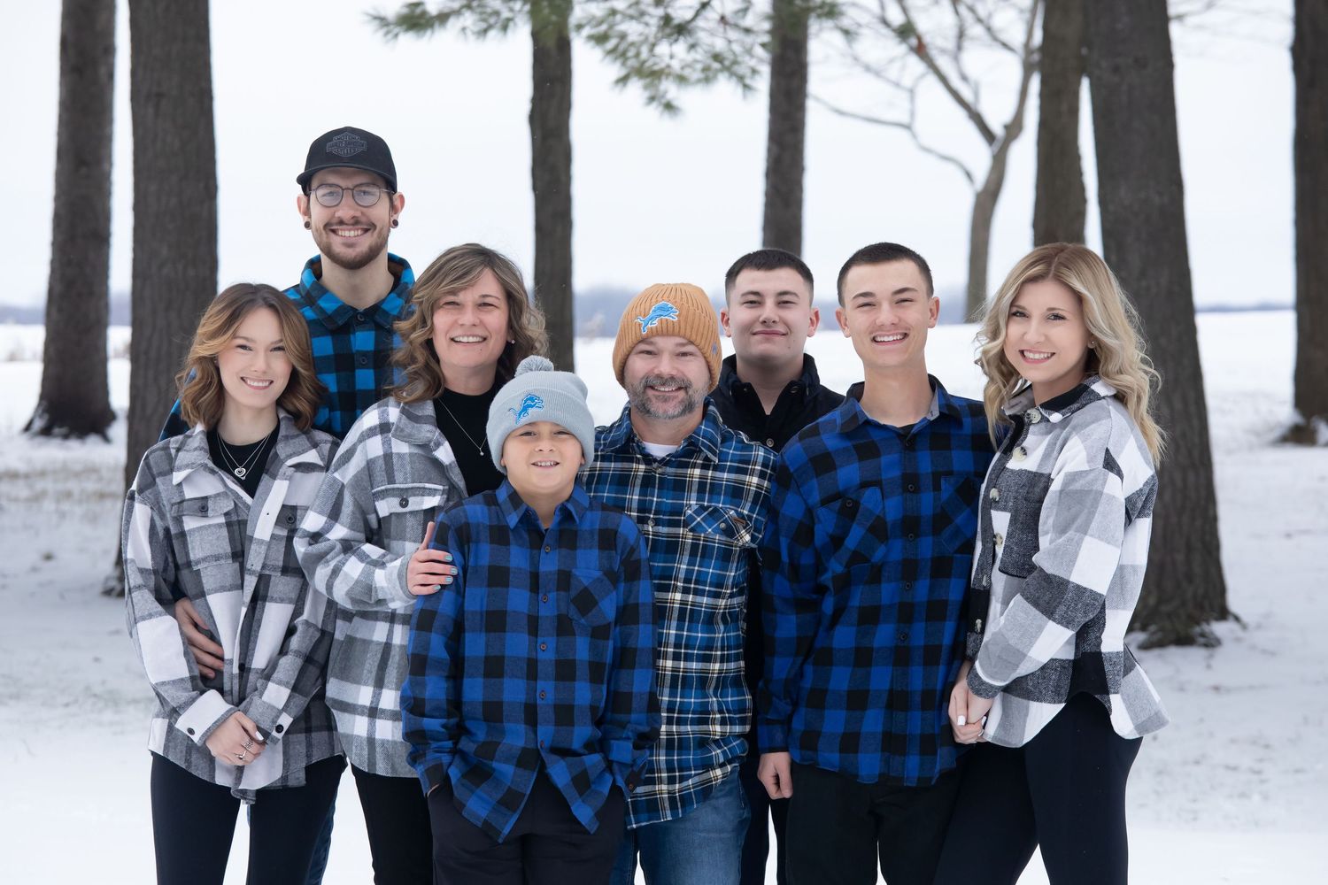 Family of eight poses in matching blue plaid flannel shirts for a winter portrait among snow-covered trees.