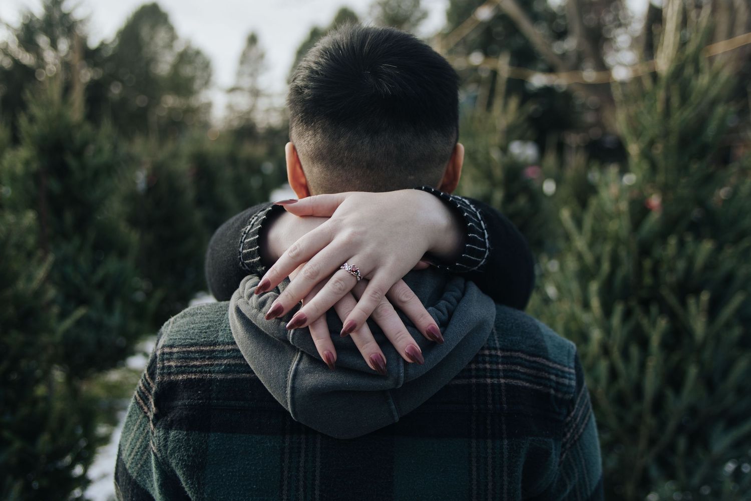 A close-up view of hands embracing on shoulders with evergreen trees in background.