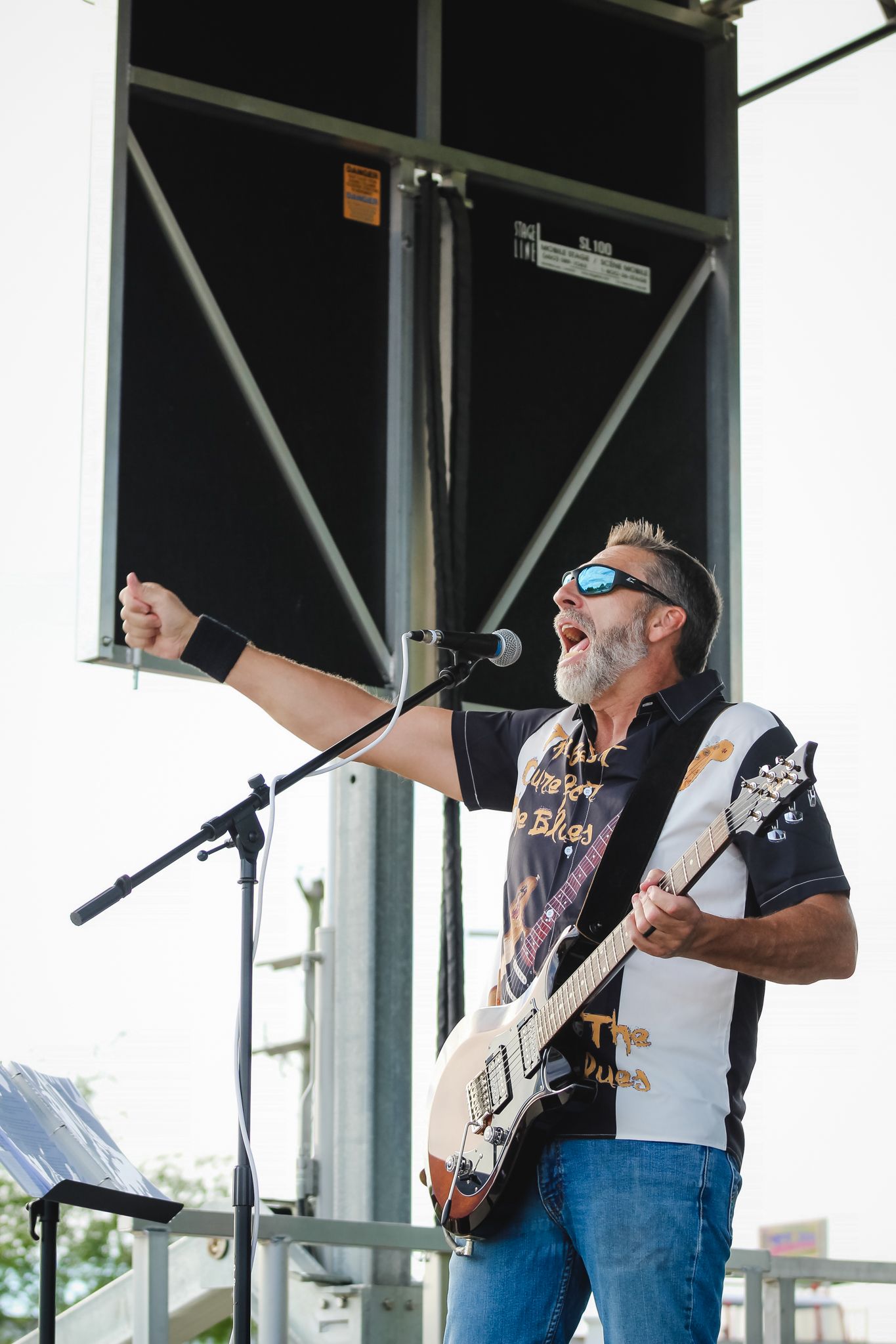 Male guitarist singing passionately on outdoor stage, wearing sunglasses and blues band t-shirt, performing at music festival.