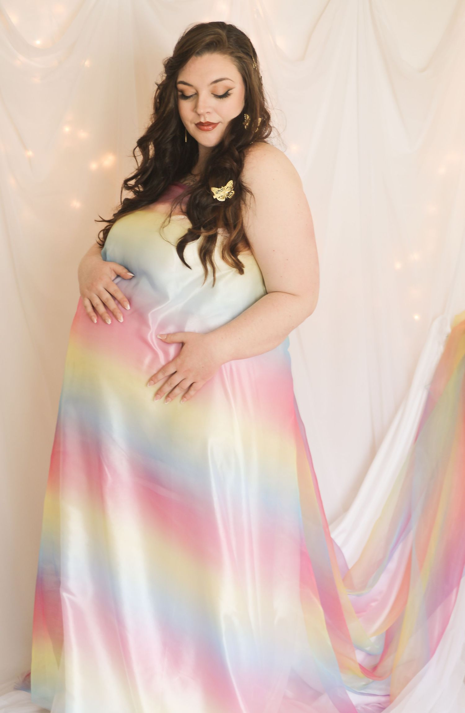 Pregnant woman in rainbow ombre maternity gown with butterfly hair clip, posing against draped white fabric backdrop.